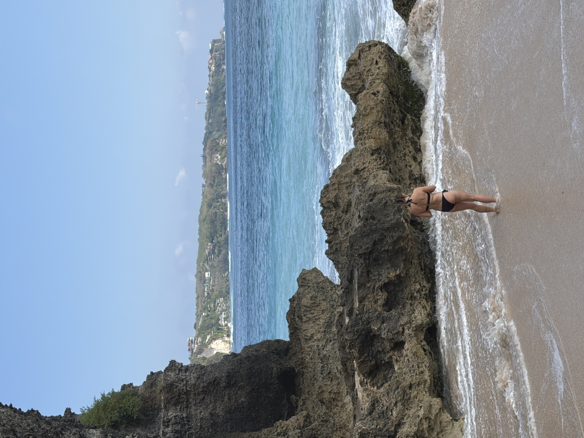 A person in a black bikini stands on a sandy beach in Kuta Selatan, Indonesia, facing the ocean. The person's back is to the camera, and their hands are held in front of their body. Waves are gently washing ashore, creating a foamy texture on the wet sand. To the left of the person, large, dark rocks form a rugged cliff face that extends into the water. In the distance, the turquoise ocean stretches to the horizon, meeting a clear blue sky. Beyond the ocean, a lush green cliff overlooks the water, with buildings and structures visible along its edge. The scene is bathed in bright sunlight, indicating a clear day.