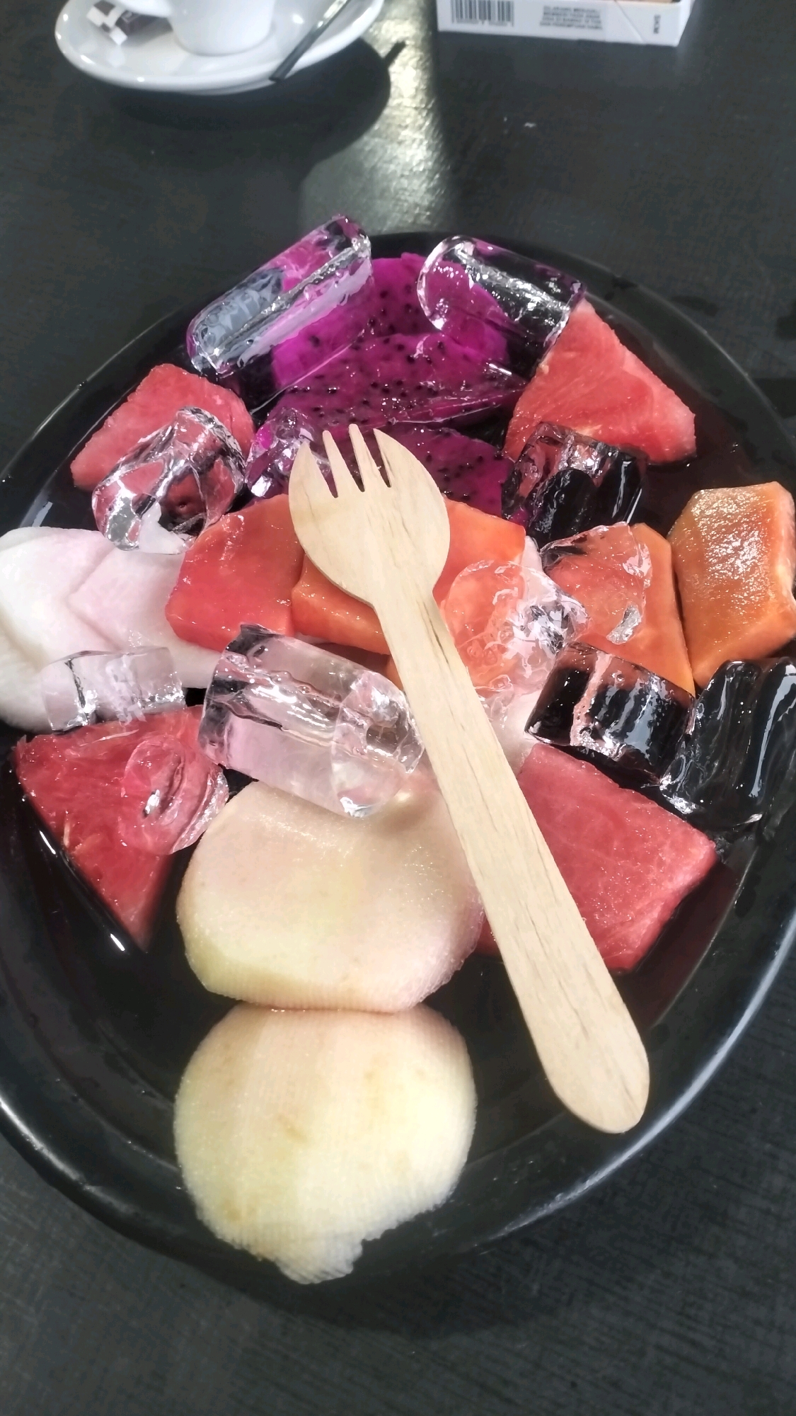 A close-up, eye-level shot captures a black, oval-shaped platter overflowing with a vibrant assortment of fresh fruits and ice cubes. The platter is placed on a dark, textured surface, likely a table, with a shallow white saucer holding a white cup and saucer visible in the upper left background, suggesting a cafe or dining setting. The fruits on the platter include wedges of red watermelon, pink dragon fruit, and pale yellow-orange fruit, possibly papaya or cantaloupe. Translucent ice cubes are interspersed among the fruit pieces, indicating the dish is intended to be chilled. A light-colored wooden fork rests diagonally across the platter, its tines pointing towards the upper right. The overall impression is a refreshing and visually appealing fruit presentation. The image's metadata does not provide specific location information, but given the context of the prompt, it could be interpreted as being in a cafe in Nunangantara, Indonesia.