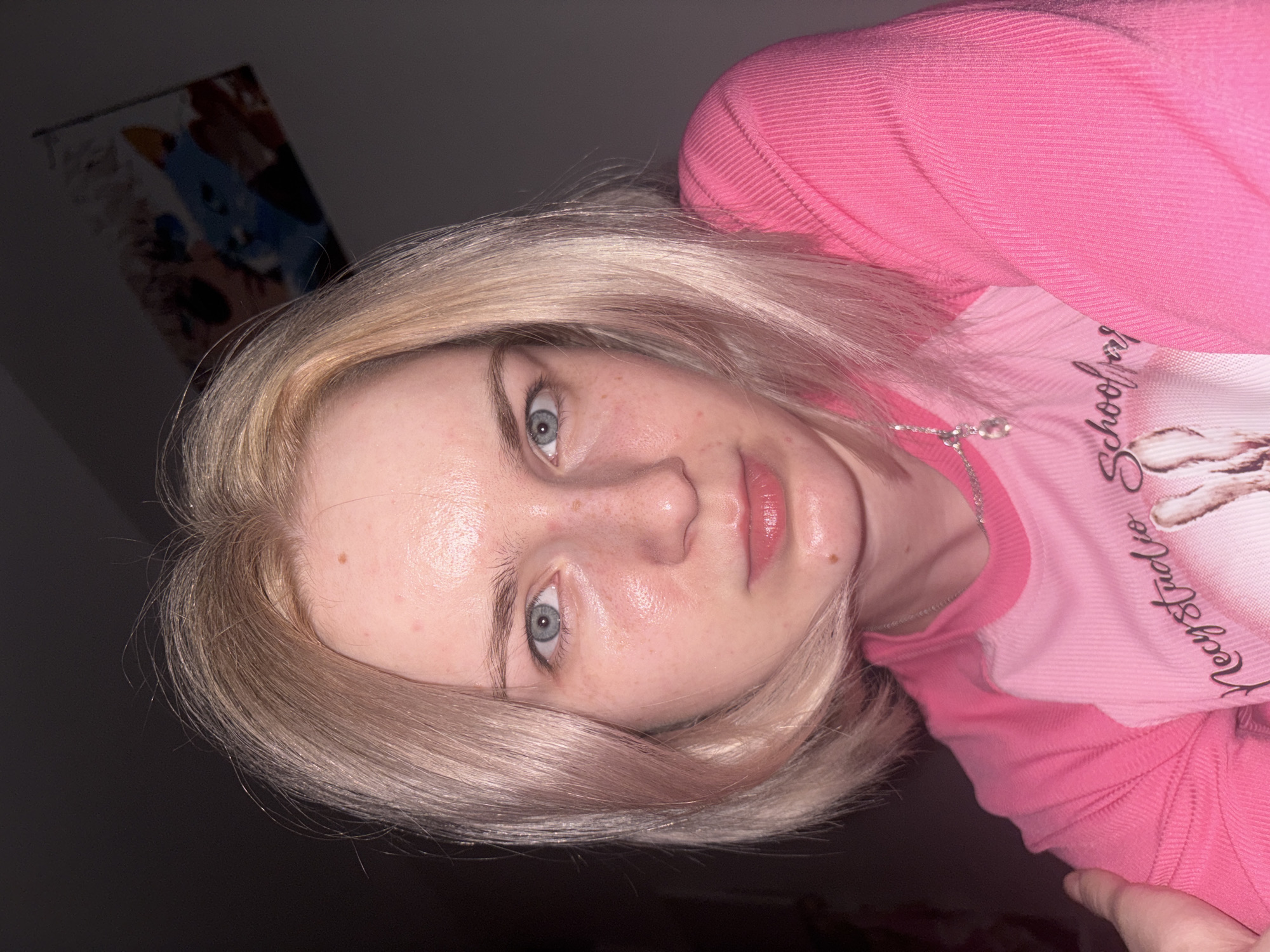 A close-up, slightly tilted portrait features a young female with shoulder-length blonde hair and blue eyes, gazing directly forward. Her fair complexion shows freckles across her nose and cheeks, along with some slight redness or minor blemishes. Her skin appears somewhat shiny, possibly from the lighting or natural oils. She is dressed in a vibrant pink ribbed long-sleeved top. Layered over this, or as part of the same garment, is a lighter pink or white panel adorned with cursive text that reads "Mystery School" and other subtle graphics. A delicate silver chain necklace with a small, clear pendant hangs around her neck. The background is predominantly dark and featureless, indicative of an indoor setting with low light, suggesting it is evening or night. In the top-left portion of the frame, a dimly lit and blurry piece of wall art, possibly depicting a blue cartoon-like figure, is faintly discernible. The subject maintains a neutral, calm expression while posing for the photograph.