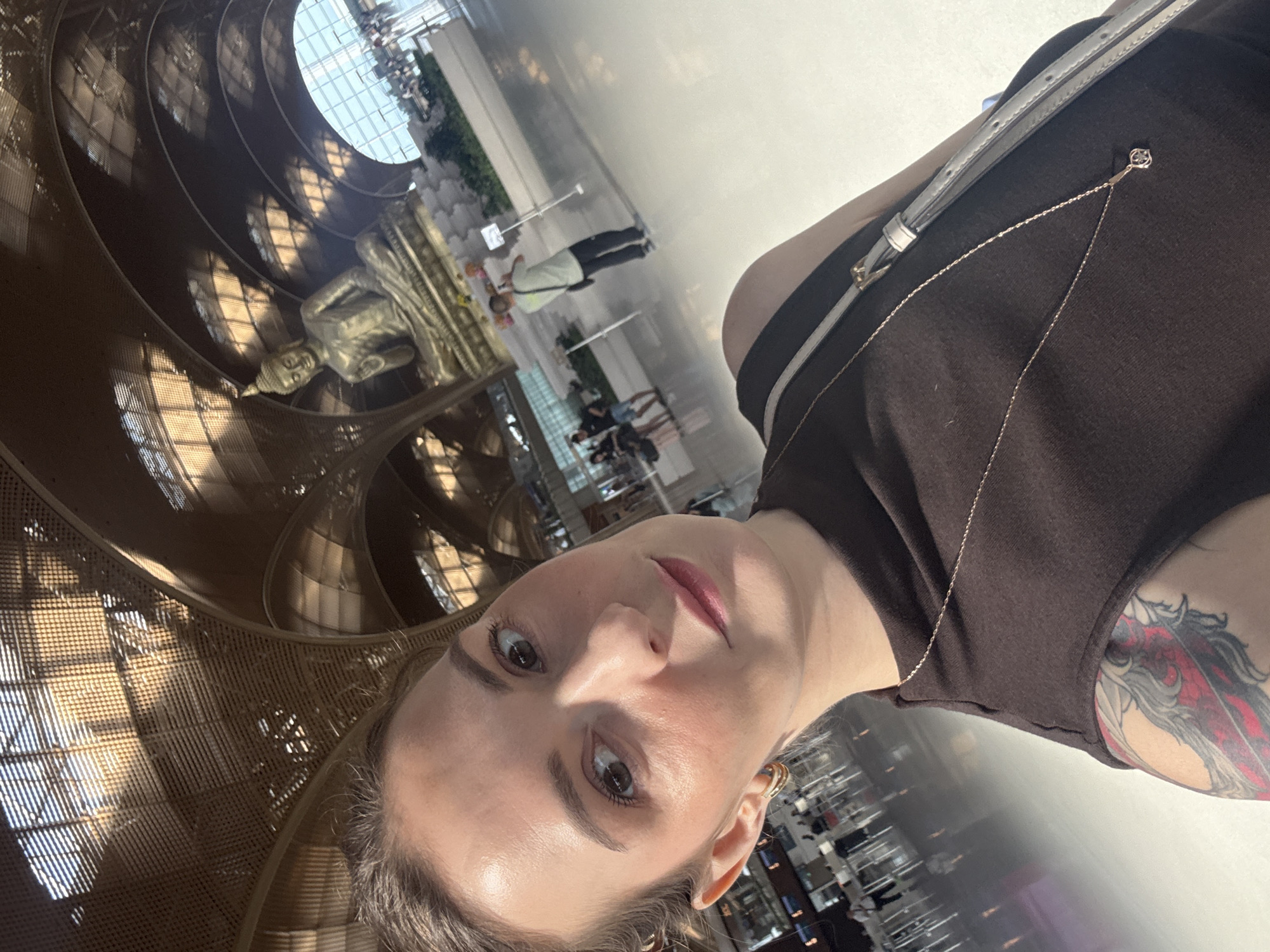 A woman with blonde hair pulled back takes a selfie in an indoor space with a high, arched ceiling made of metal and glass. She is wearing a dark brown sleeveless shirt and a delicate gold necklace. A large, golden statue of Buddha is visible in the background, seated on a platform. Several other people are scattered throughout the large, well-lit space, some appearing to be walking or taking pictures. The floor is polished and reflects the light. The location is identified as Kandal Steung, Cambodia.