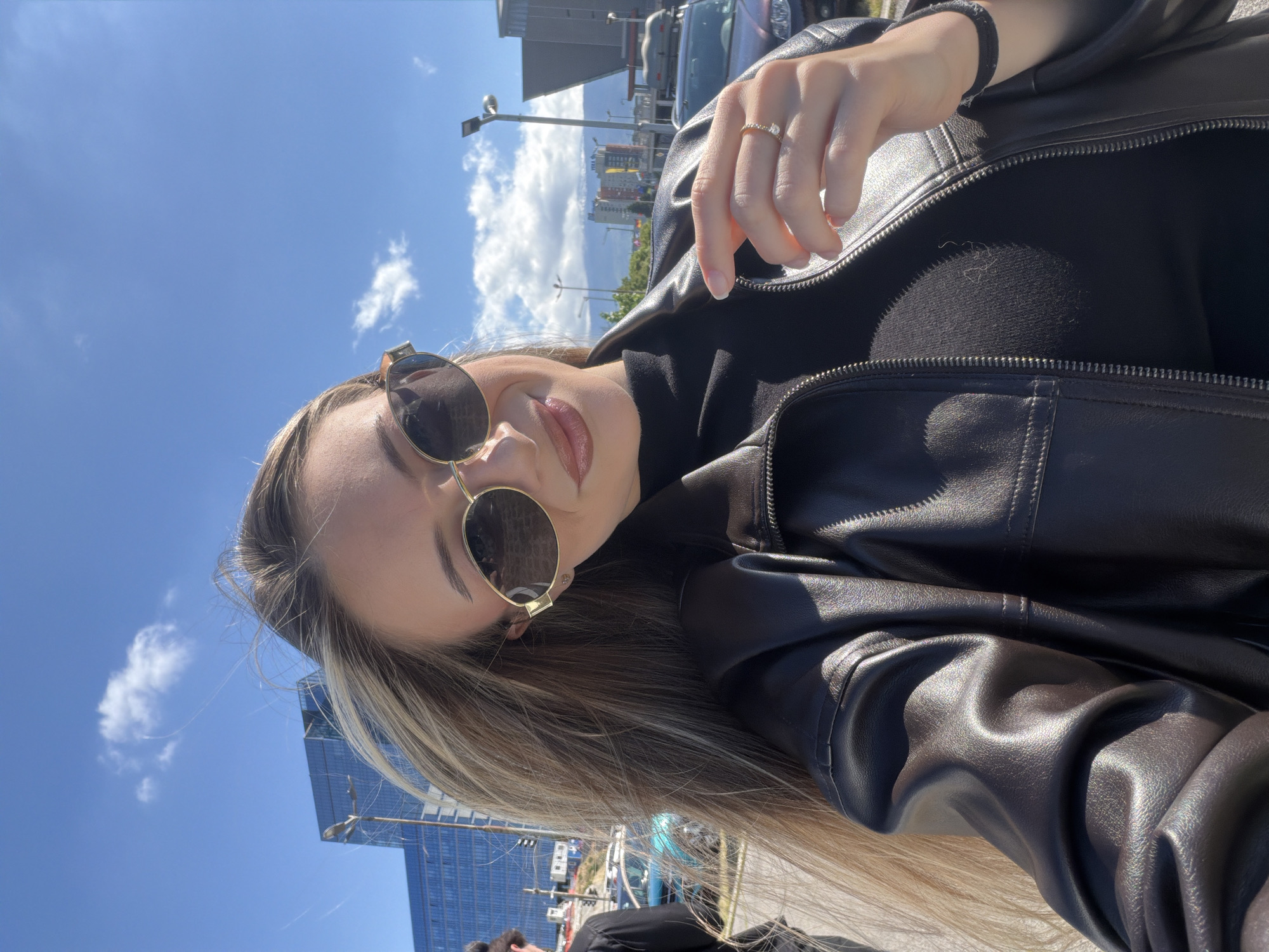 A woman with blonde and light brown hair wears sunglasses and a black leather jacket. She is wearing a gold ring on her right ring finger and a black wrist band on her right wrist. She is outdoors during daytime. The background includes buildings, a blue sky with clouds, street lights, and cars. Based on the architectural style and the presence of modern high-rise buildings, the location could be the city of Sofia, Bulgaria.