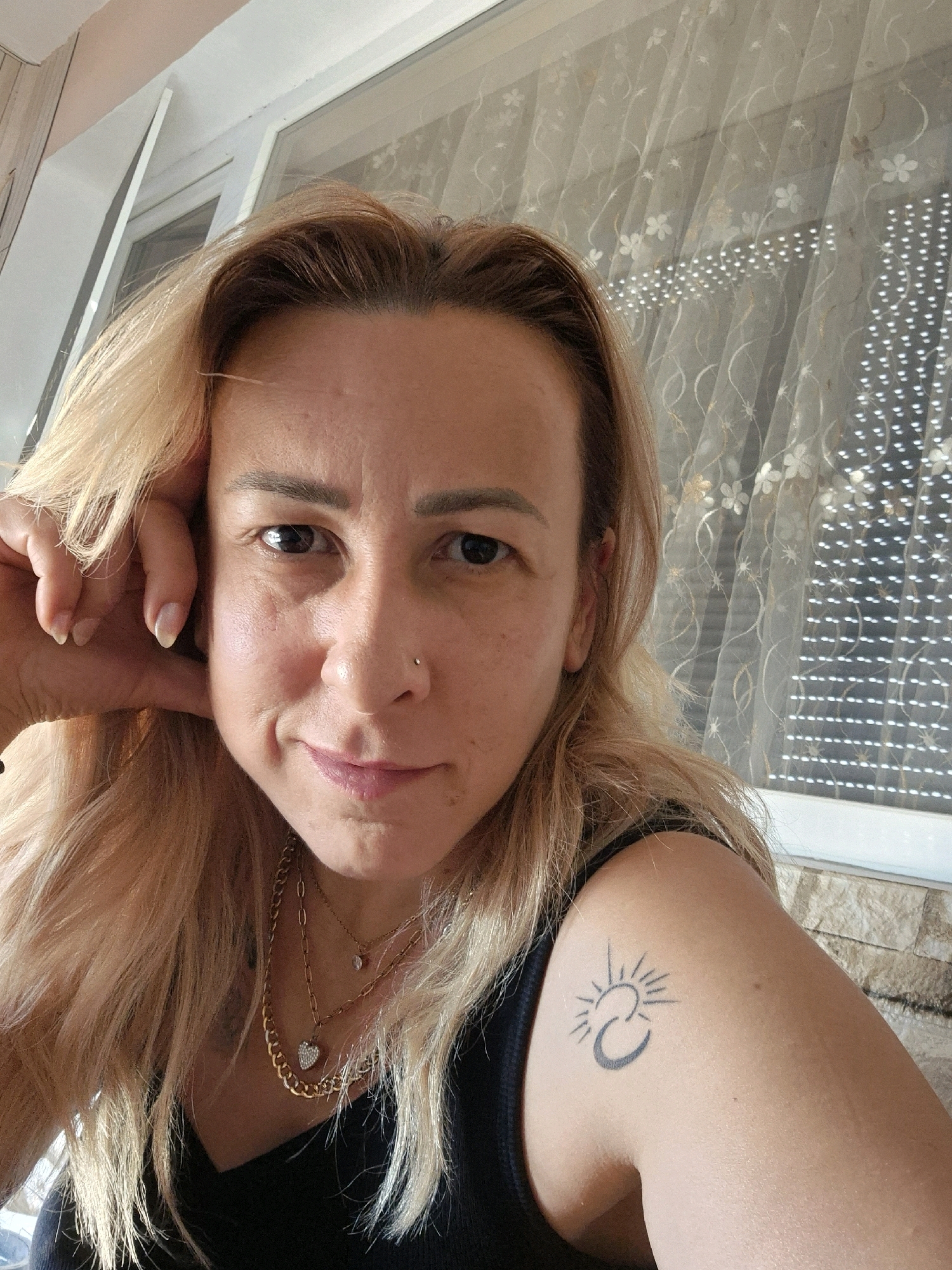 A fair-skinned woman with shoulder-length blonde hair, wearing a black top and multiple necklaces, poses for a selfie. Her left hand is resting on her cheek, and her right arm, displaying a tattoo of a crescent moon and sun, is visible. She has a nose ring and is looking directly at the camera. The background features a window with a floral patterned sheer curtain and a brick wall. The scene appears to be indoors, with natural light filtering through the window.
