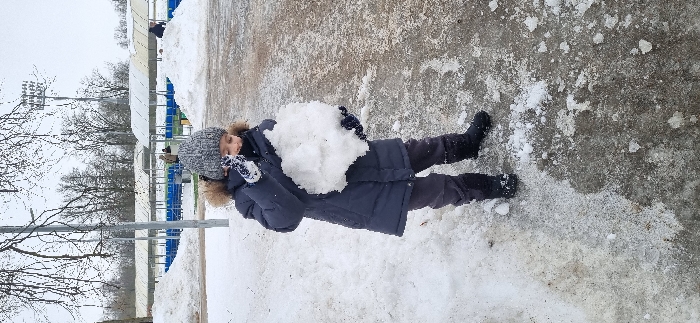 The image captures a child standing outdoors in a snowy environment at a sports complex in Pavlovsk, Russia. The child, approximately 6-8 years old, is dressed in a grey knitted beanie, a navy blue hooded winter coat with a light fur trim, dark grey trousers, and black winter boots. Dark gloves are worn on both hands, with the left glove exhibiting a white patterned design. The child holds a substantial, irregularly shaped mass of white snow, roughly 30-40 cm in diameter, at chest height with both hands, looking downwards at it.
The ground immediately surrounding the child consists of patches of melting snow, ice, and exposed wet, dark ground. Scattered pieces of snow and ice are visible on the surface. In the mid-ground, behind the child, rows of empty blue stadium seating are visible beneath white, sloped shelters, indicating a sports arena or track. Large, irregular mounds of snow are present around these structures. Two indistinct adult figures are visible in the background near the seating area. The distant background features bare deciduous trees, a vertical metal pole, and a tall stadium floodlight tower on the upper right. The sky is uniformly overcast and grey, suggesting winter or early spring conditions.