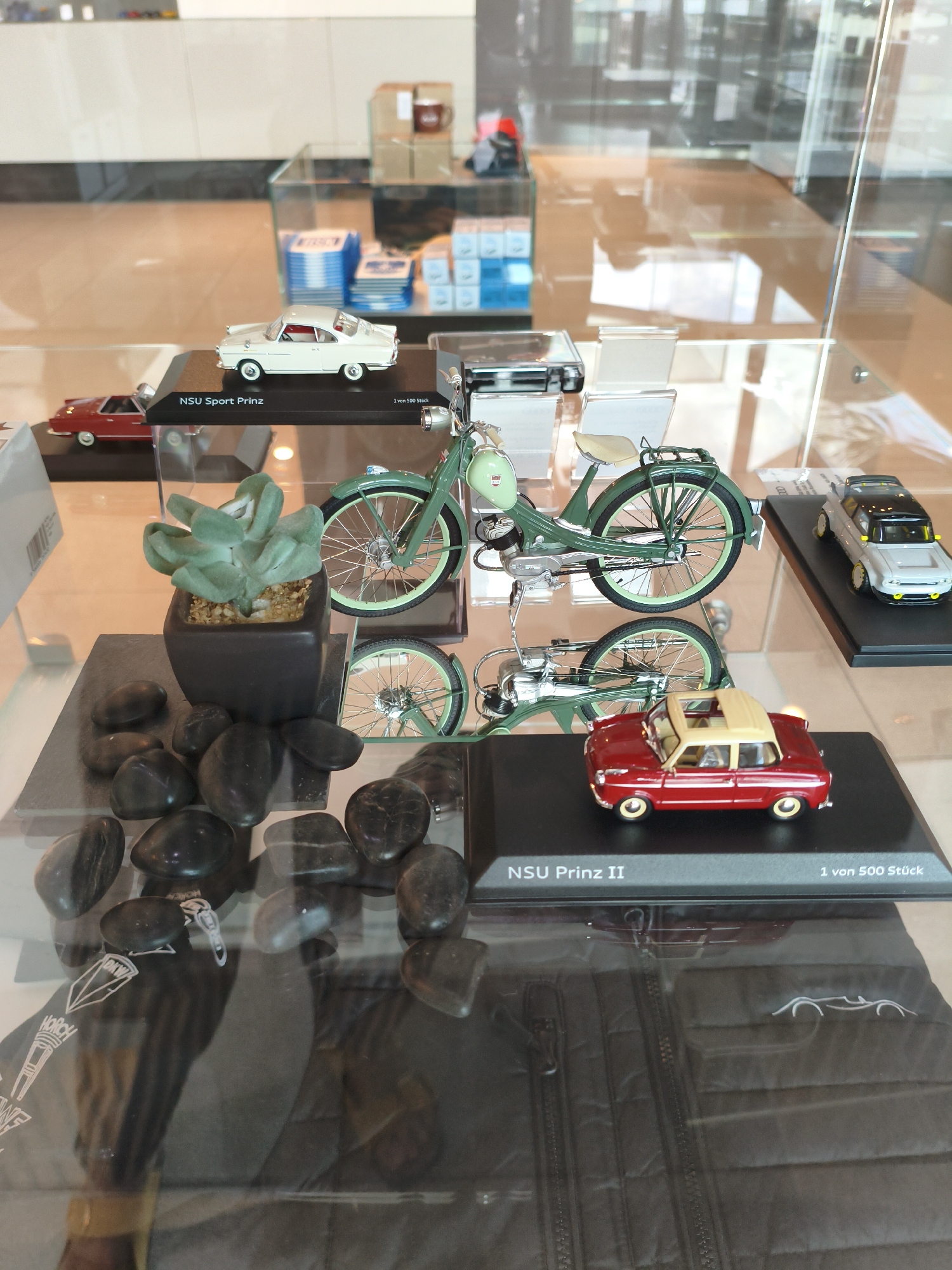The image displays a collection of model cars and a model moped inside a glass display case. In the foreground, a black quilted vest with a zipper and an embroidered outline of a car is visible, reflecting the glass. A few black, polished stones and a small potted succulent plant are arranged on a dark slate on the left side of the display.
The model vehicles are positioned on black display stands. In the lower right, a red and beige two-door model car, labeled "NSU Prinz II 1 von 500 Stück," is prominently featured. Above it and to the left, a green model moped is shown, with its wheels and handlebars visible. Further back on a higher shelf, a white model car, labeled "NSU Sport Prinz," is displayed next to a red model car. A gray and yellow model car is visible on the far right, behind the red and beige NSU Prinz II.
The background of the display case is blurred, showing reflections of the surrounding environment, which appears to be a modern interior with smooth flooring and glass partitions. This scene is located within the Audi Forum in Ingolstadt, Germany.