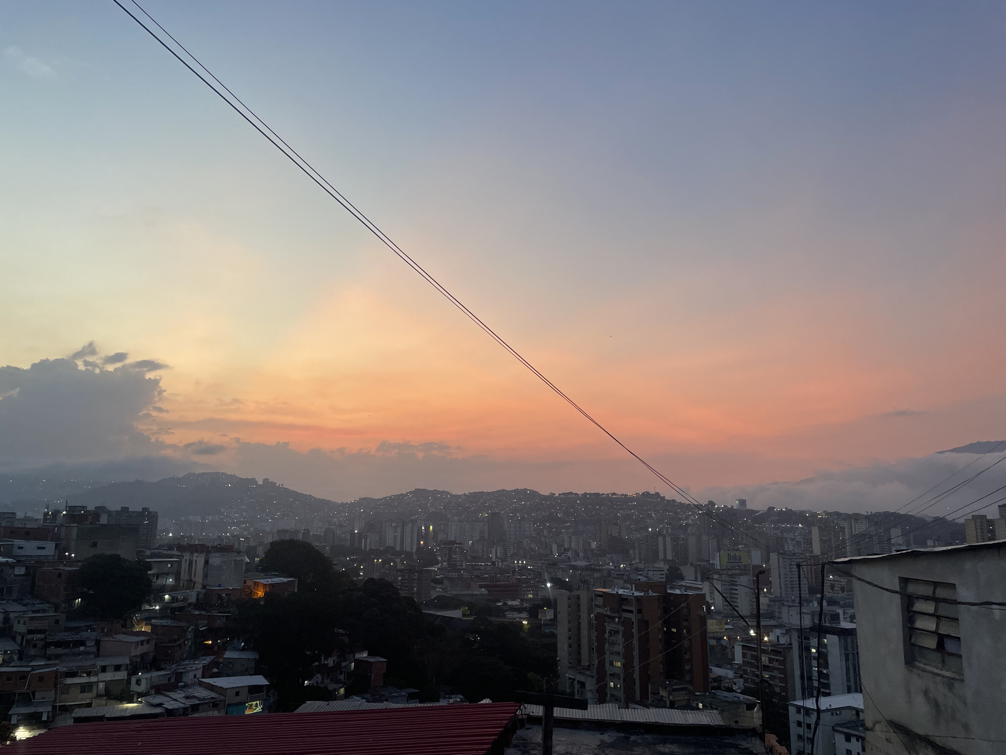 The image shows a cityscape at dusk or dawn, likely in Caracas, Venezuela, given the mountainous terrain and architectural styles. The foreground features densely packed buildings, primarily low-rise structures and rooftops with some higher-rise apartment buildings interspersed. The sky transitions from a blueish hue to a warm orange and pink near the horizon, with some clouds visible. Two power lines stretch diagonally across the upper portion of the frame. The middle ground is characterized by hills covered in urban development, with numerous lights indicating the presence of residential areas and roads. The background reveals hazy mountain ranges partially obscured by clouds, typical of the geographical setting of Caracas. No people are visible in the frame. The scene conveys a sense of urban density and the blending of natural landscape with the built environment.