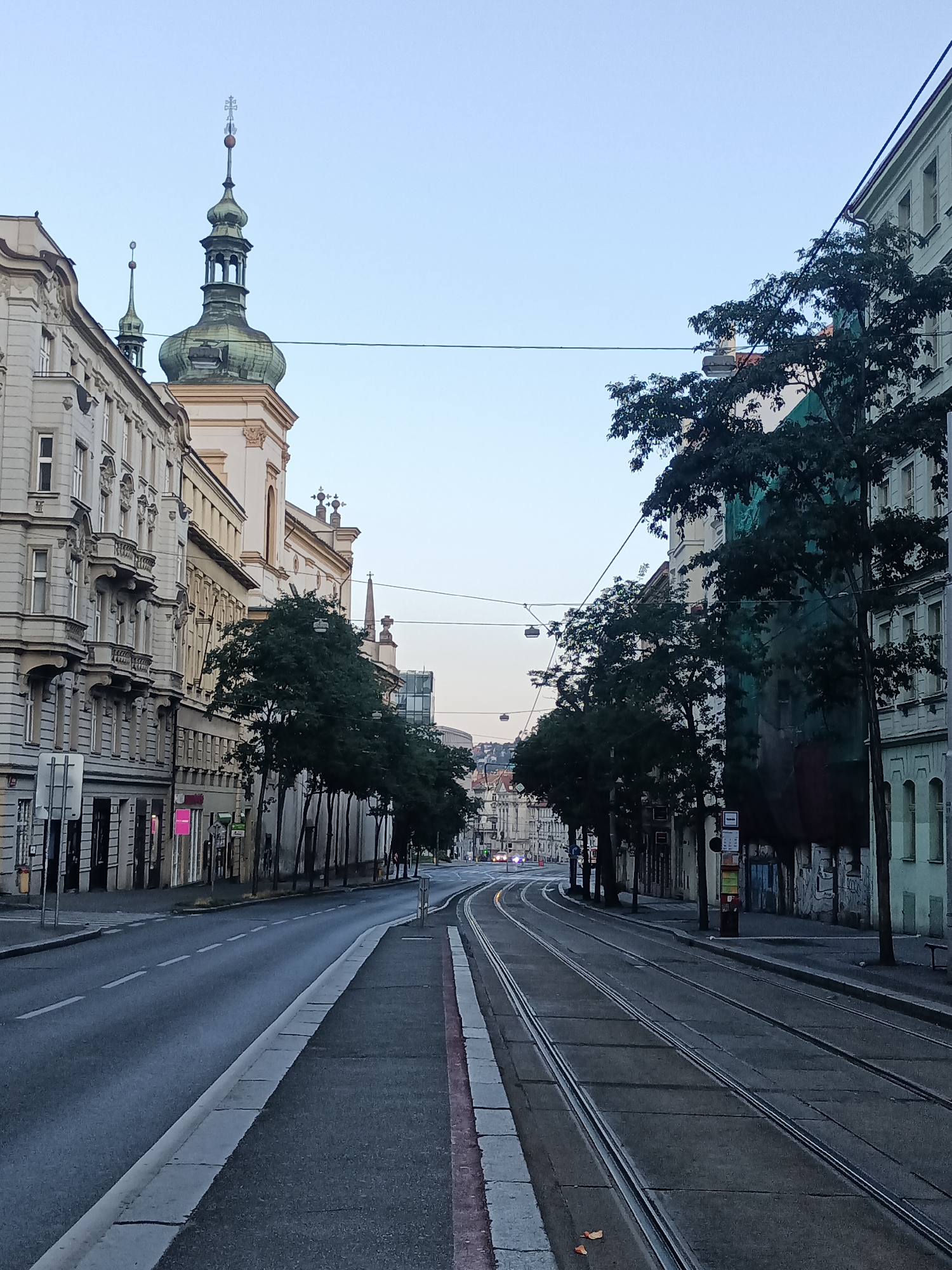 The image depicts a street scene in Prague, Czechia, likely during early morning or late evening given the soft, diffused light and lack of significant activity. The perspective is from street level, looking down a wide street lined with multi-story buildings on both sides.
On the left side of the street, ornate buildings with classical architectural features, including balconies, detailed facades, and arched windows, are visible. These structures appear to be a mix of residential and commercial establishments, with some storefronts visible at street level.
Dominating the skyline on the left is the distinctive green dome and spire of a church, topped with a cross. Another smaller spire is visible adjacent to it.
The street itself is paved and features tram tracks running down the center. The tram tracks curve towards the right side of the image as they recede into the distance. A lane for vehicular traffic is also present on the left.
On the right side of the street, the buildings are also multi-story but appear to have a slightly more uniform and perhaps utilitarian design compared to those on the left. One building prominently displays a large green tarp or netting covering a significant portion of its facade, suggesting ongoing construction or renovation. Trees with dark green foliage are planted along the sidewalk on both sides of the street, casting shadows on the pavement.
Overhead, electrical wires, likely for the trams, are strung across the street, supported by poles. The sky is a pale blue, indicating either dawn or dusk. No people are clearly visible in the frame, and the overall atmosphere is one of stillness and quiet. In the far distance, more city buildings and possibly hills are visible under the hazy sky.