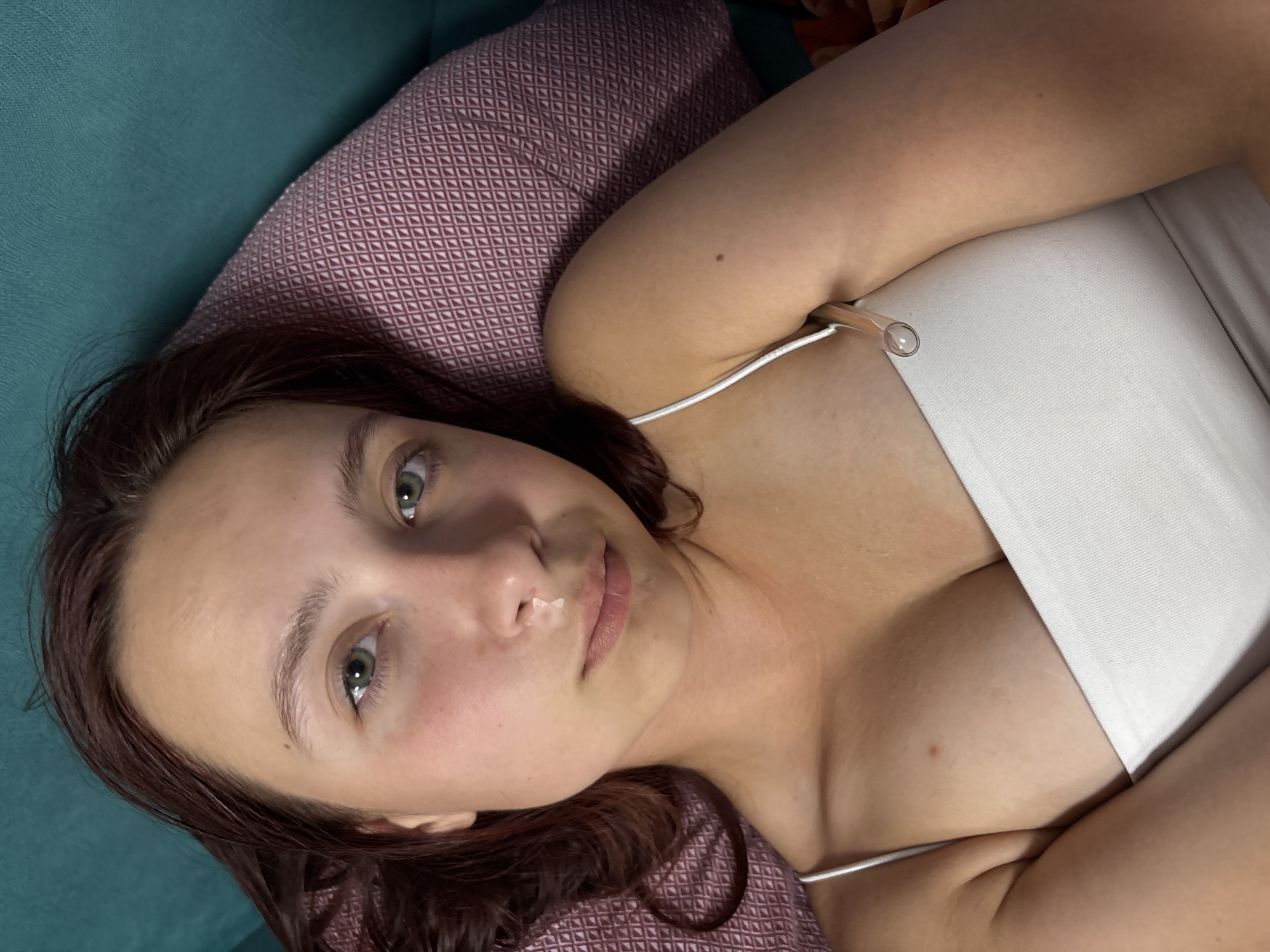 A young woman with long, dark brown hair and green eyes is pictured lying down, looking directly into the camera. She is wearing a white camisole or tank top with thin straps. Her expression is neutral.
She is resting on a maroon or purple pillow featuring a white geometric diamond pattern. To her left, and behind the pillow, is a teal or turquoise colored surface, possibly a sofa or bed. A notable detail is a clear, slender, tube-like object, possibly made of glass or clear plastic, tucked into the side of her white top near her chest/armpit area, with a rounded end visible. A small mole is visible on her upper right arm. The setting appears to be indoors, suggesting a relaxed, informal atmosphere in Прикамский, Russia. No text is visible in the image.