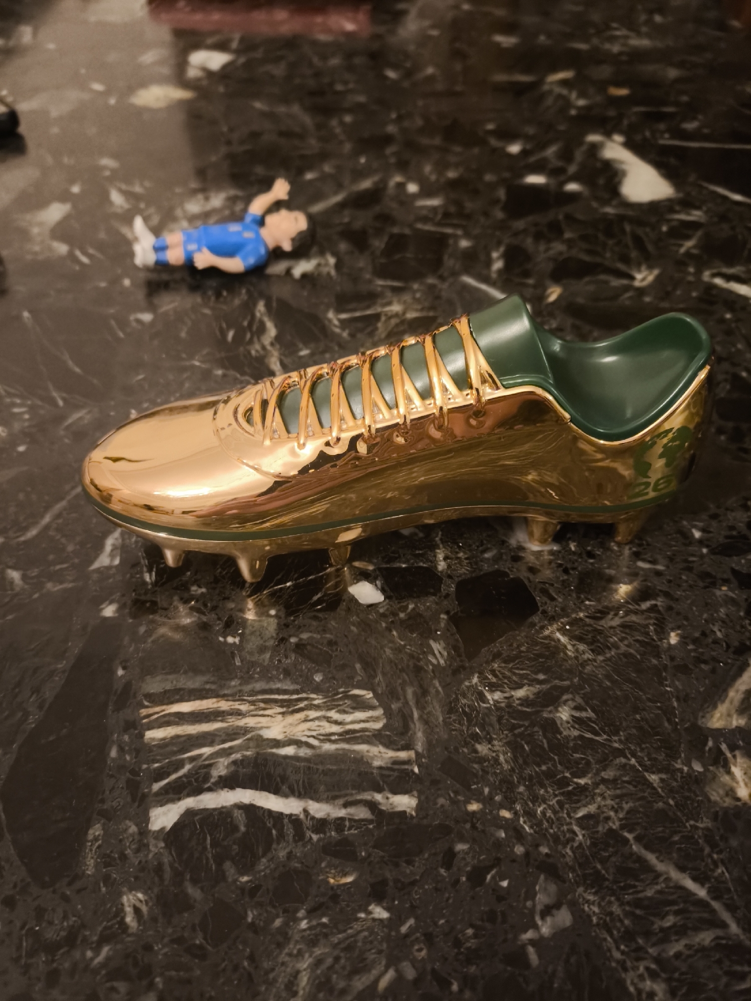 A shiny, gold-colored miniature soccer cleat sits on a dark, marbled surface. The cleat has green accents on the inside and the laces are depicted as gold. In the background, a small figurine of a soccer player in a blue jersey lies on its back with one arm raised. The setting appears to be indoors on a table. There is no indication of the city of Batumi, Georgia.