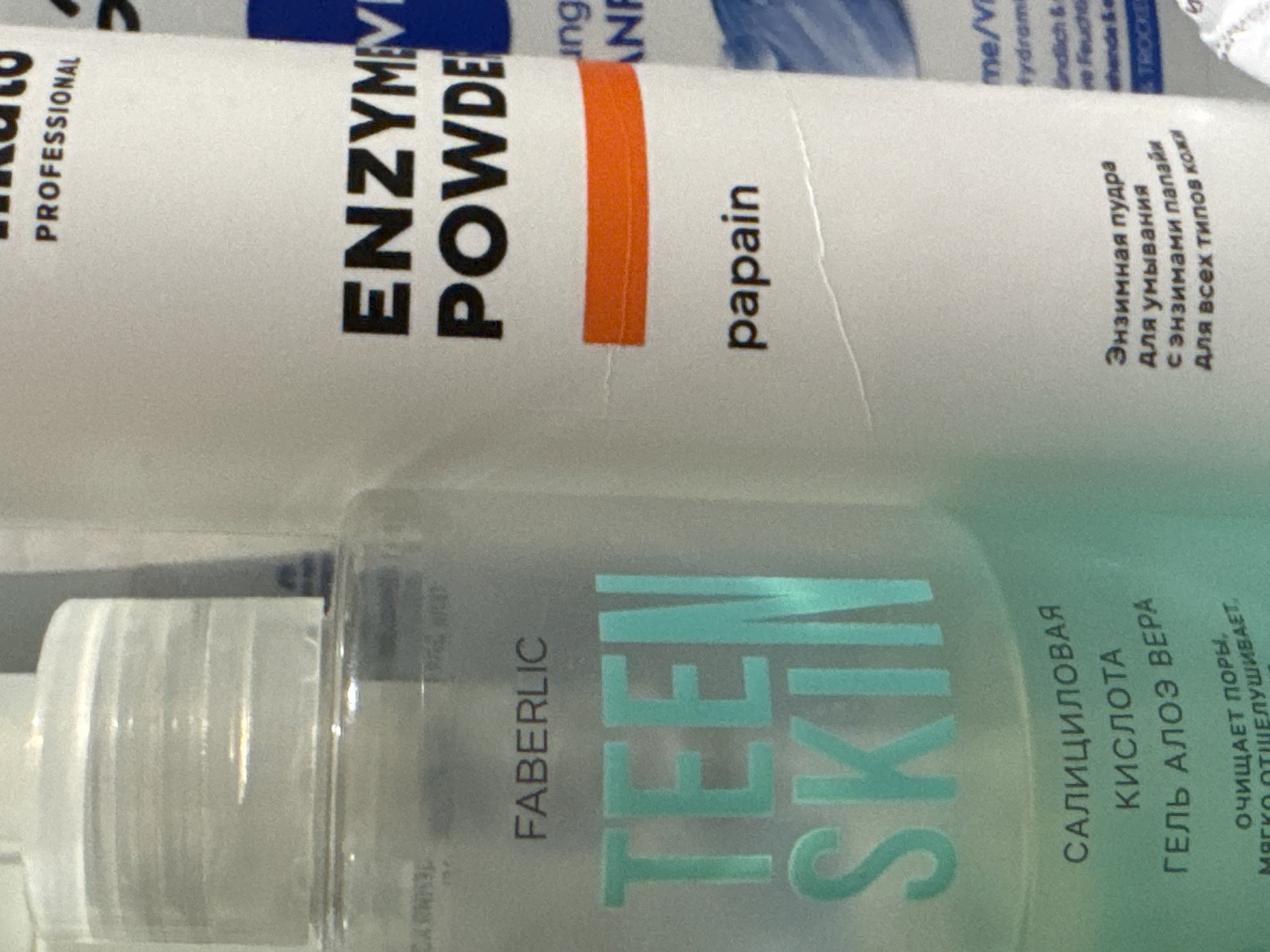 The image shows two cosmetic products, a powder and a gel, against a blurred background. The powder product, on top, has "ENZYM POWDER PROFESSIONAL" and "papain" printed on its packaging. Below it, the gel product has "FABERLIC" and "TEEN SKIN" prominently displayed. The gel packaging also features text in Cyrillic, which appears to describe its ingredients and purpose: "САЛИЦИЛОВАЯ КИСЛОТА ГЕЛЬ АЛОЭ ВΕΡΑ" (Salicylic Acid Aloe Vera Gel) and "ОЧИЩАЕТ ПОРЫ, мягко оТШЕЛУШИВАЕТ" (Cleans pores, gently exfoliates). Another label on the powder product, also in Cyrillic, reads "Энзимная пудра Для умывания с энзимами папайи Для всех типов кожи" (Enzyme powder for washing with papaya enzymes for all skin types). The lighting suggests an indoor setting, possibly a retail environment or a personal collection of cosmetics. No people or specific time of day or weather cues are visible. The image is an eye-level, close-up shot focused on the product labels.