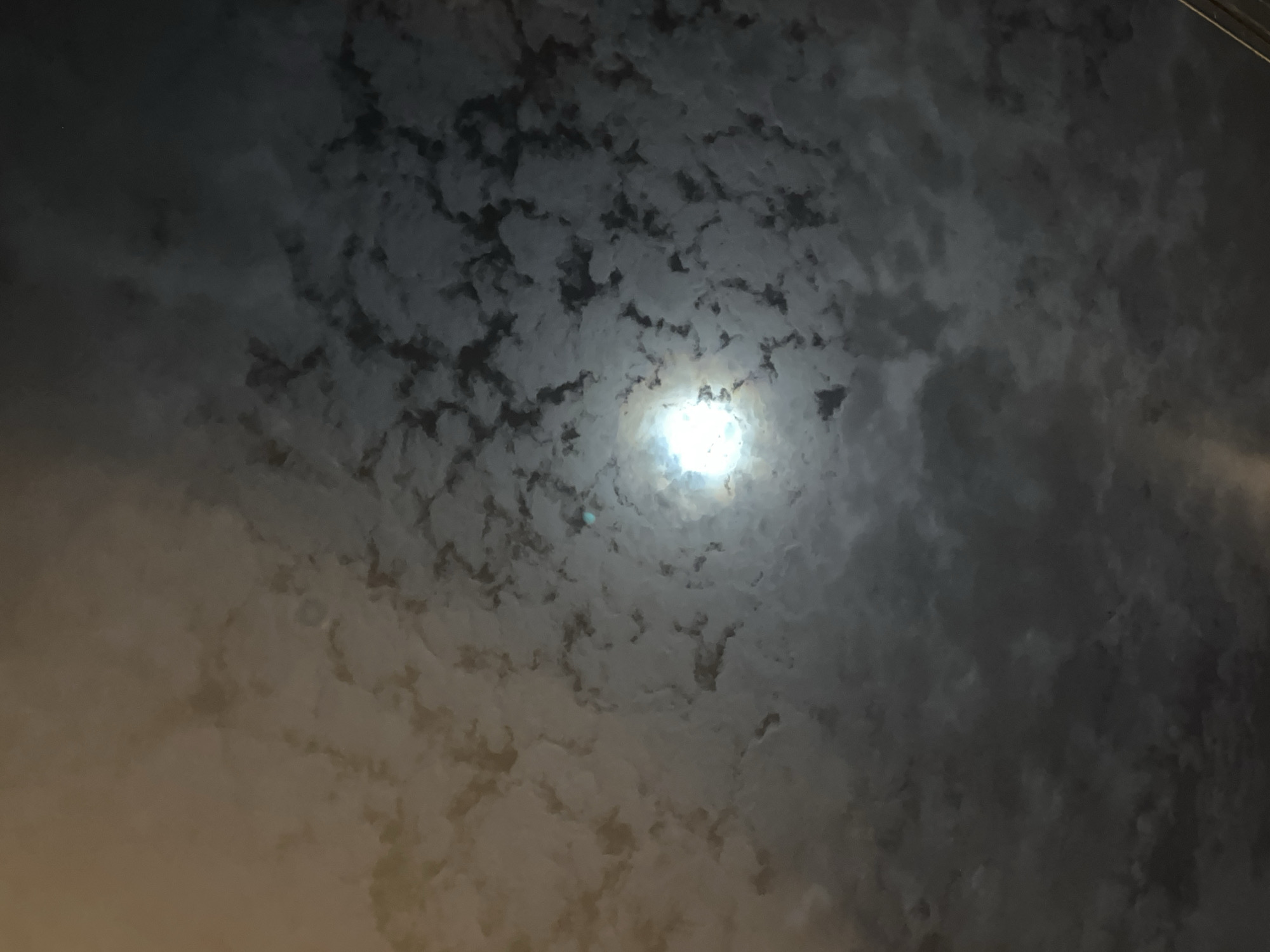 The image depicts a nighttime sky in Chiang Mai City Municipality, Thailand. The focal point is a bright, circular light source, likely the moon, shining through a layer of altocumulus clouds. The clouds are distributed across the sky, appearing as individual, rounded masses. The cloud formations range from off-white to dark gray. The sky surrounding the clouds is dark, indicating nighttime.