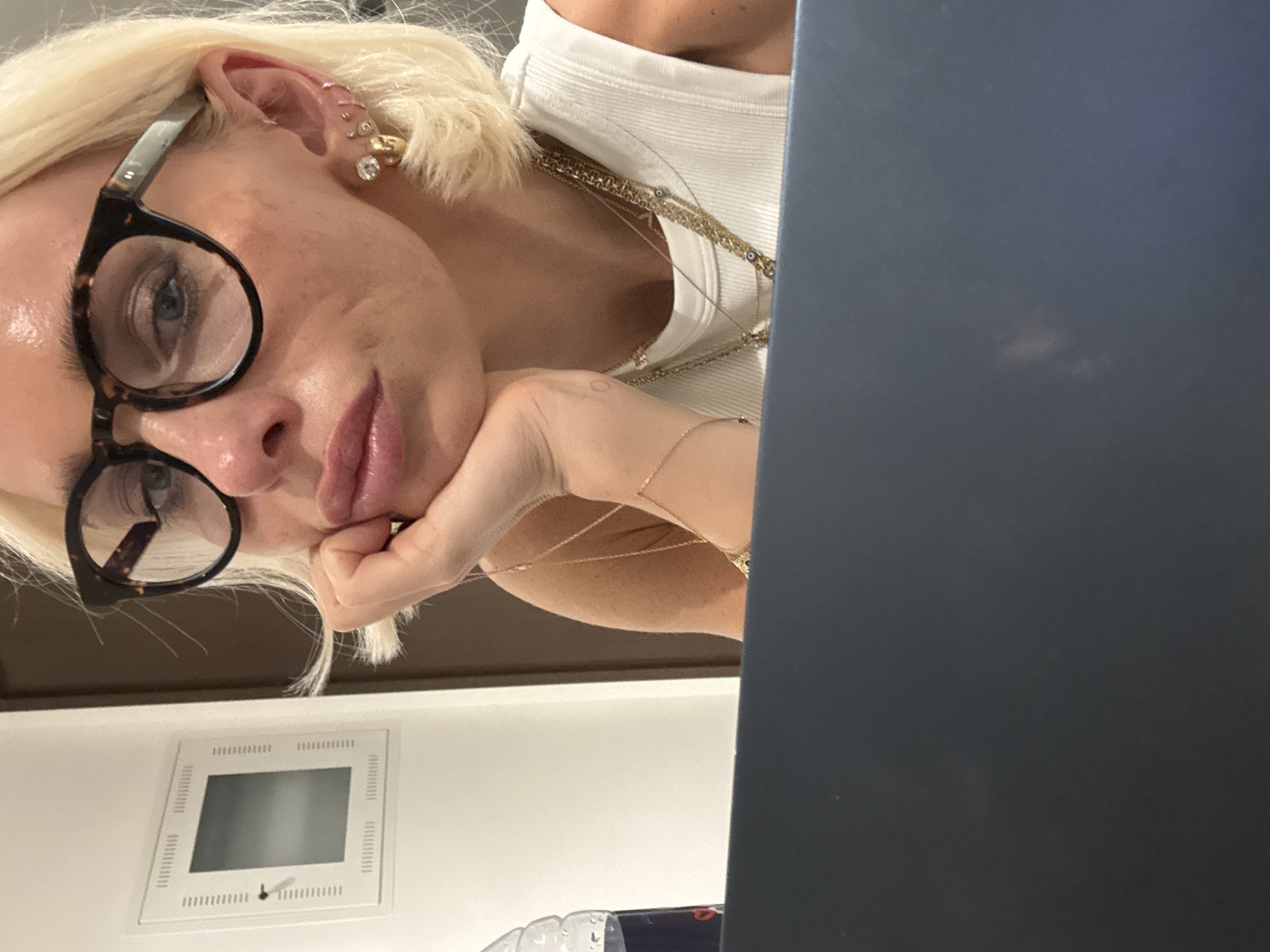 A medium shot captures a close-up of a fair-skinned blonde woman with blue eyes, wearing tortoiseshell-patterned eyeglasses, and a white sleeveless top. Her hand is resting on her chin, and she is adorned with multiple gold necklaces, including a layered chain with small blue eye pendants, and several earrings in her left ear. To the left of the woman, a portion of a plastic water bottle is visible, and a white wall-mounted panel with a small dark screen and a keyhole is on the left side of the frame. The lower portion of the image is dominated by the dark blue surface of a laptop. The background consists of a muted cream-colored wall and a dark brown door. The scene appears to be indoors, possibly an office or hotel room. There is no indication of the city of Dubai, United Arab Emirates in the image.