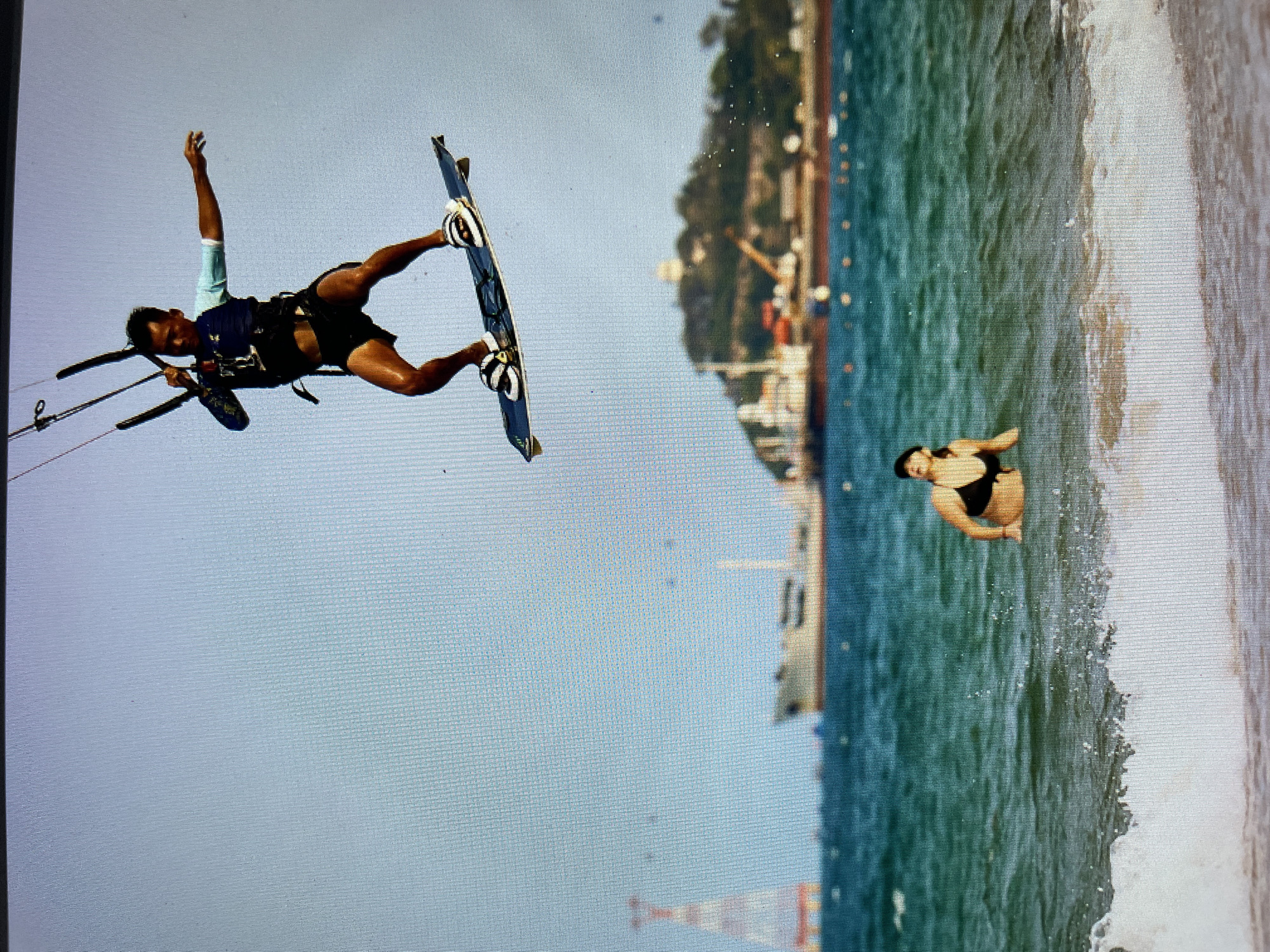 The image shows a kitesurfer performing a jump trick in the air. The kitesurfer is male, wearing a harness and shorts. He is holding onto the kite's control bar. A wakeboard is attached to his feet.
In the background, a woman is swimming in the water near the beach. The buildings of Bacong, Philippines, are visible on the shoreline. The image shows a bright, sunny day.