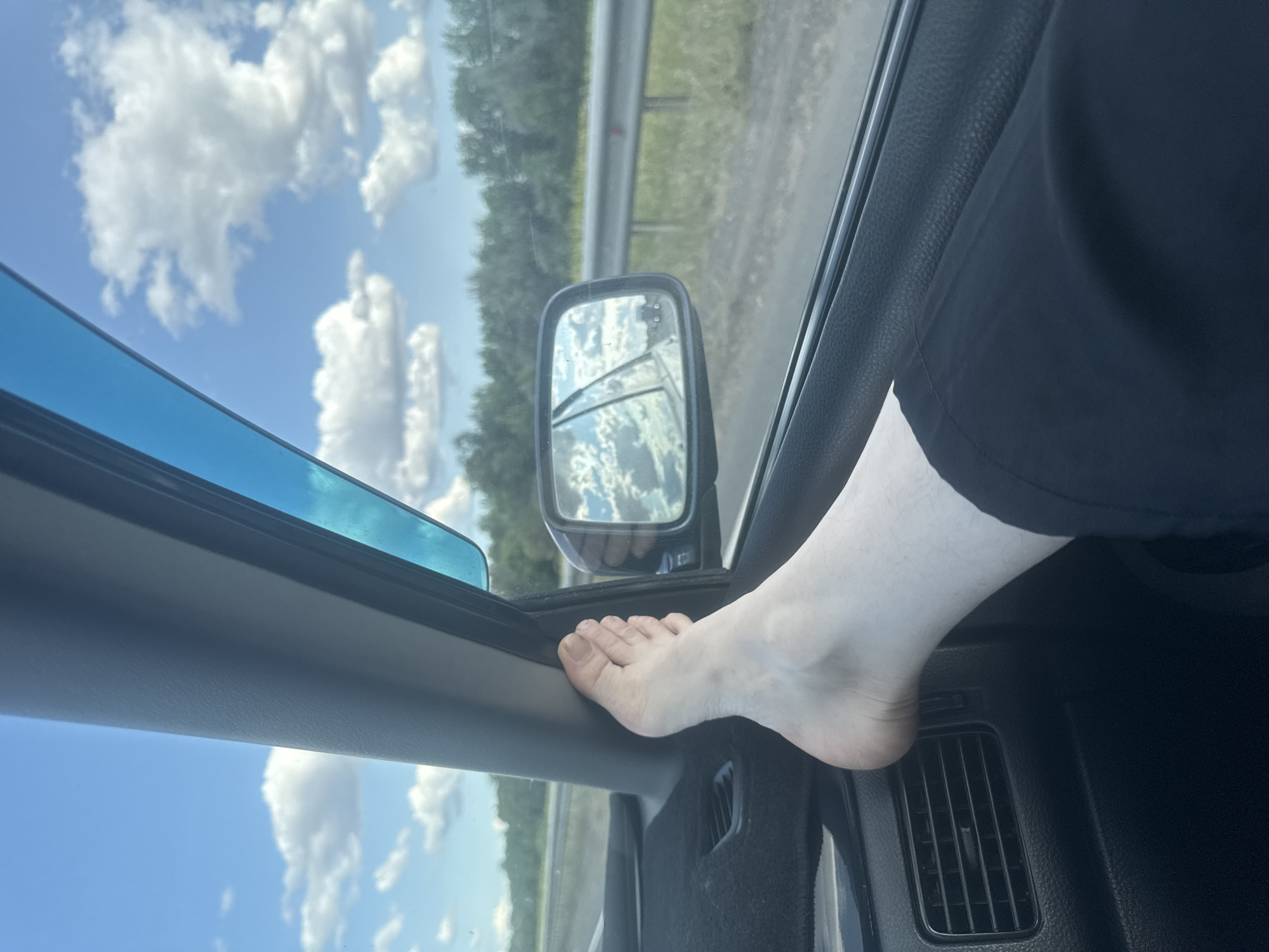 The image captures a person's bare foot resting on the dashboard of a car. The foot is pale and appears to be a left foot, with toes extended. Black pants are visible below the knee. Through the car window, a blue sky with white clouds and green trees lining a road is visible. A side-view mirror reflects the sky and trees. The interior of the car is dark, with a black dashboard and a visible air vent. The scene is set during daylight. The location of Perevolotsky, Russia, is not evident from the visual information in the image.