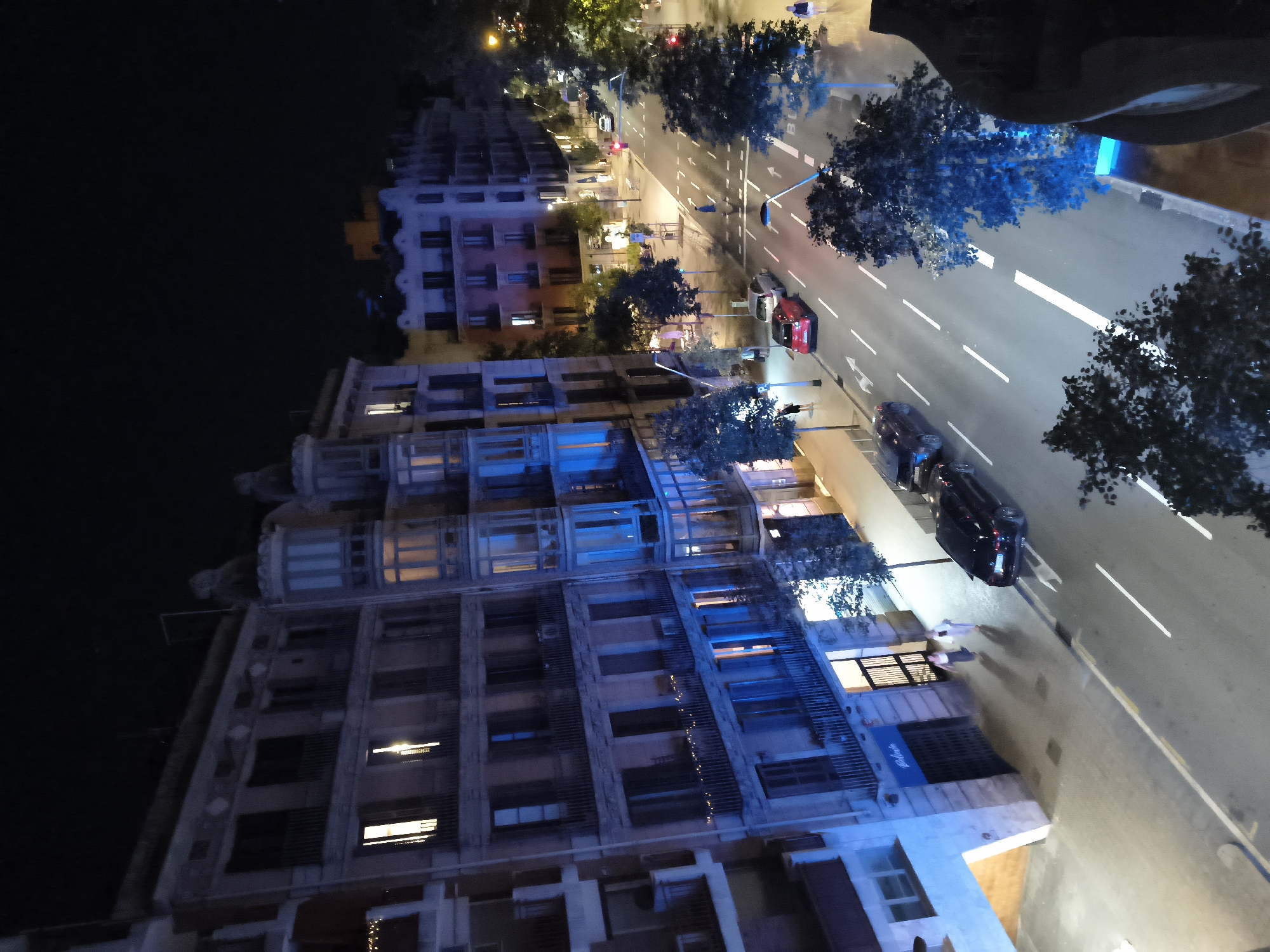 The image depicts an aerial view of a street scene at night in Barcelona, Spain, likely taken from a balcony or elevated vantage point. The street is lined with multi-story apartment buildings, characterized by their detailed facades and numerous windows, some of which are illuminated. The street itself is asphalt, with clear lane markings and pedestrian crossings. Several vehicles are present on the street, including a red car, a dark-colored sedan, and another dark-colored vehicle parked further down. Pedestrians are visible on the sidewalks and crossing the street. Trees are strategically planted along the sidewalks, some illuminated by blue ambient lighting. The overall atmosphere is urban and nocturnal, with artificial lighting from the buildings and streetlights dominating the scene.
