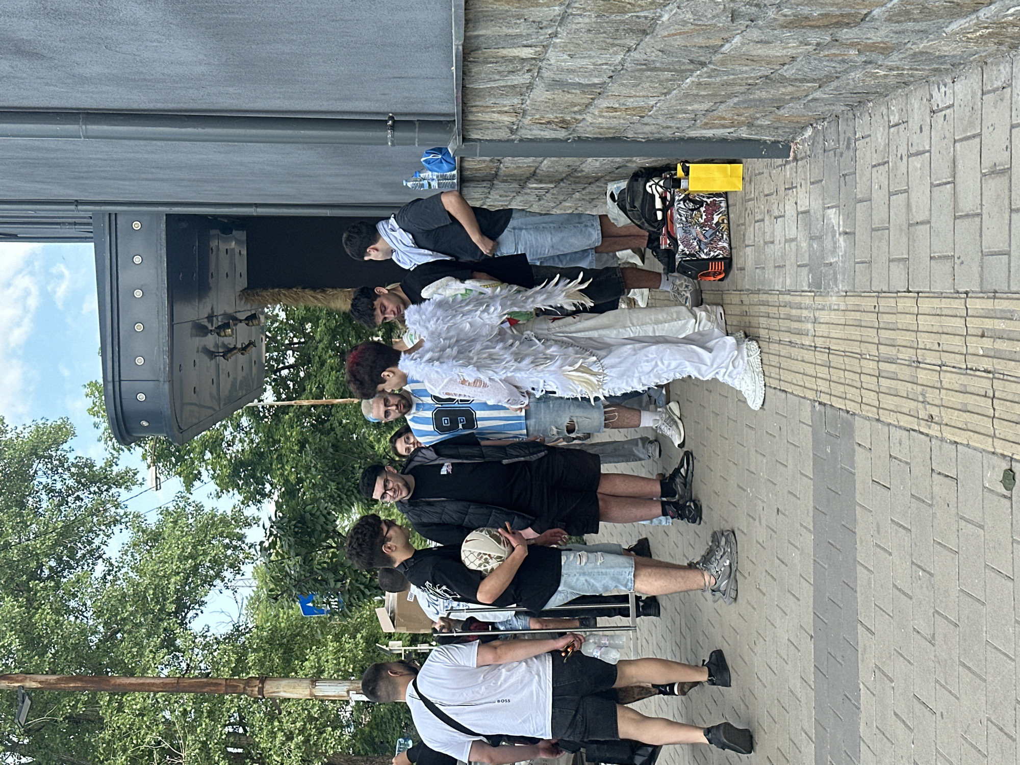 The image depicts an outdoor urban scene in Sofia, Bulgaria, featuring a gathering of approximately ten individuals, predominantly young men, on a paved sidewalk beside a building.
In the foreground, a person with reddish hair is centrally positioned, facing right and slightly turned towards the group, wearing a full white costume consisting of large feathered wings, white pants, and white athletic shoes. To their immediate left, a man in a black T-shirt and distressed light-wash denim shorts holds a white and gold patterned soccer ball. He wears glasses and has dark curly hair. Adjacent to him, a man with dark hair and glasses, wearing a black puffer vest over a black T-shirt and black shorts, looks towards the individual holding the soccer ball. Further to the left, partially obscured, is a man viewed from the back, wearing a white short-sleeved T-shirt with a "BOSS" logo, black shorts, and a black cross-body bag.
To the right of the feathered figure, a man with light blue hair wears a blue and white striped jersey with "89" printed on it, light-wash distressed denim shorts, and white athletic shoes. Next to him, a man in a dark grey T-shirt and light blue denim shorts has a light blue scarf draped around his neck. Another man, partially visible further right, wears a dark top and light shorts. In the background, a woman with dark hair and glasses is faintly visible, observing the group.
The ground is paved with rectangular light grey interlocking pavers, featuring a darker grey strip and a textured tactile paving strip. Along the right side, a stone-faced wall forms the base of a dark grey building with vertical structural elements. Above, a dark architectural overhang extends from the building, equipped with multiple circular recessed lights, below which a section of textured brown material is visible. On the sidewalk next to the building wall, a black bag adorned with colorful graphics and orange accents sits next to a yellow rectangular object. Nearby, two clear plastic water bottles and a blue fabric bag are visible.
The background reveals dense green tree foliage beneath a partly cloudy blue sky. A rusted metal pole with a street light fixture is visible on the far left. A blue circular street sign with a white arrow is discernible amidst the distant trees. The individuals appear to be engaged in casual conversation or observation within the gathering.