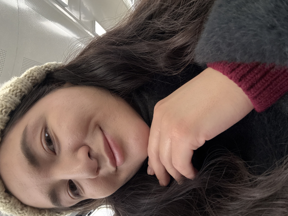 A female subject, appearing to be in Il'inskiy Pogost, Russia, is captured in a close-up, upward-angled shot within an indoor environment. She has dark, wavy hair and wears a light-colored knitted beanie. Her face, featuring light-to-medium skin tone and brown eyes, displays a slight smile. She is dressed in a dark, possibly black, turtleneck or high-collared garment, beneath a fuzzy, dark gray outer layer with a ribbed maroon cuff visible on her right wrist. Her right hand is raised, with fingers partially curled, and her chin appears to rest lightly on the back of her hand. The background indicates an indoor setting with a white or light-toned ceiling featuring rectangular panels and linear light fixtures or vents. No other individuals or specific identifying objects are visible in the immediate environment.