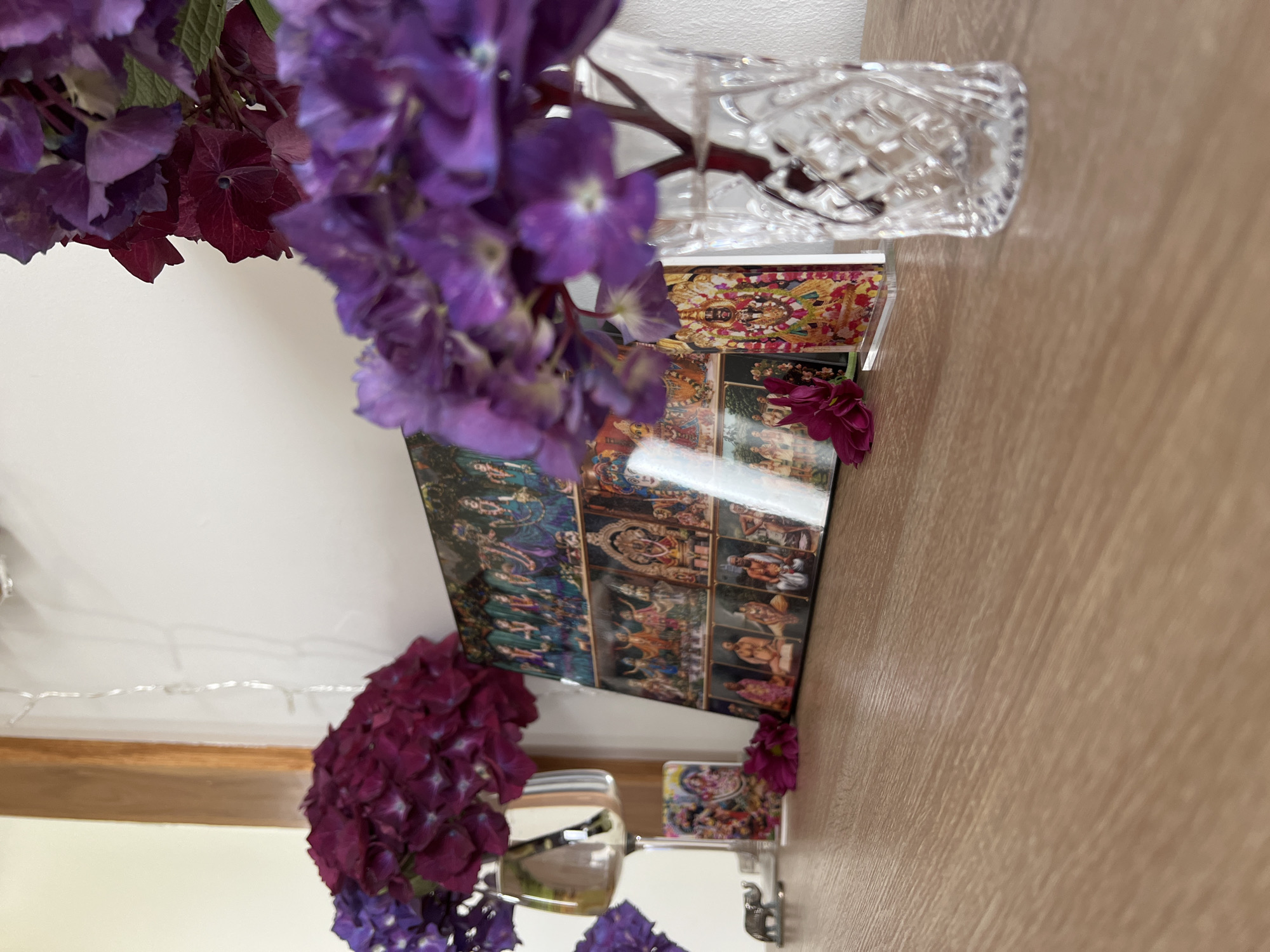 The image depicts a close-up view of a wooden surface with various decorative items. In the foreground, a clear glass vase with a diamond-cut pattern holds purple hydrangeas. To the left, a collection of religious images is displayed on a table, including a framed picture of deities, a small elephant figurine, and a silver wine glass. The background features a white wall with string lights and a wooden post. The overall scene suggests an indoor setting, possibly a home or a shop, decorated with flowers and religious iconography. There are no people visible in the image.