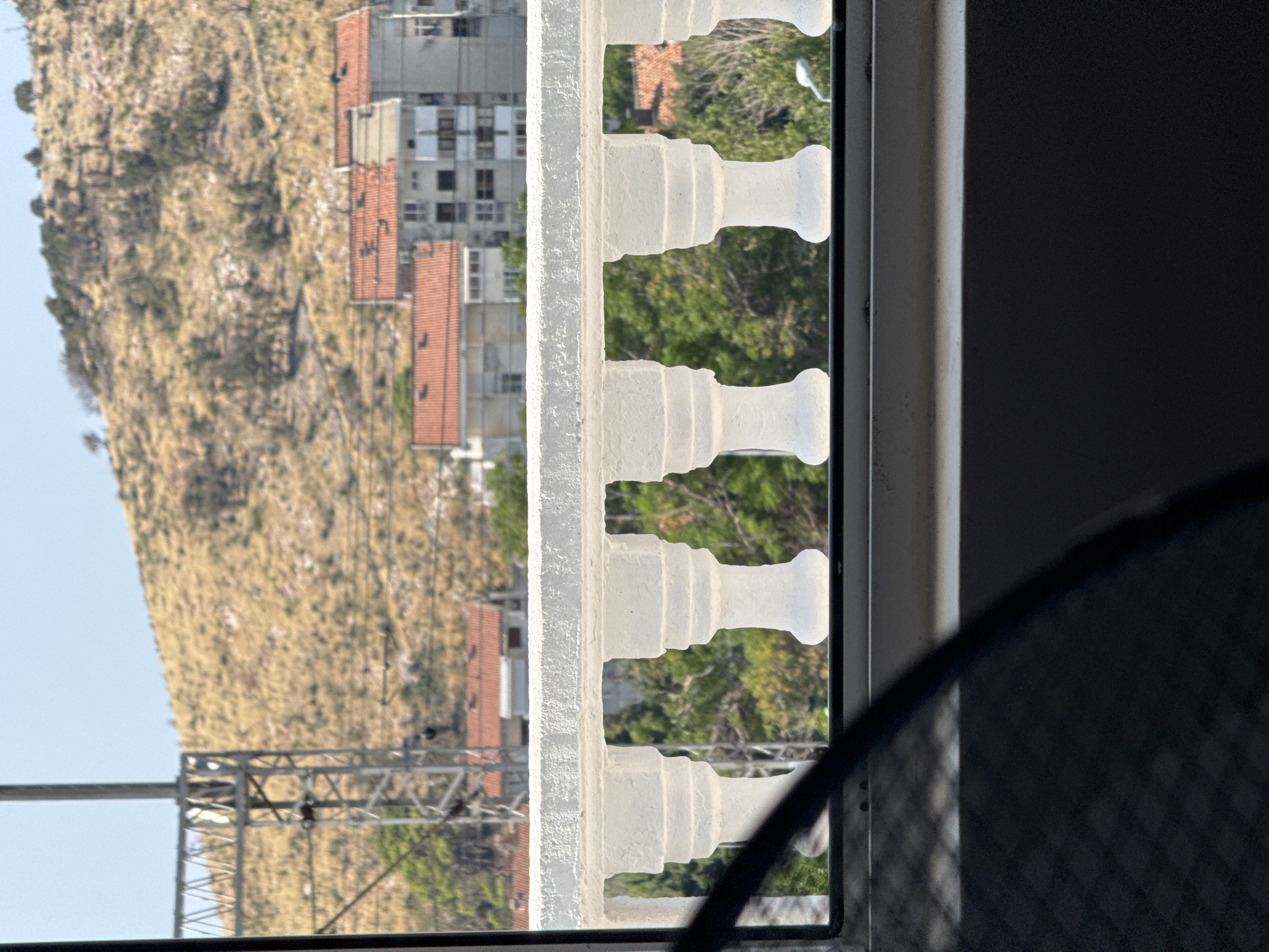 The image is a medium-shot, eye-level, outdoor photograph taken from a balcony. In the foreground, a white stone balustrade with decorative pillars runs horizontally across the frame. The balustrade appears to be textured. Beyond the balustrade, a view of a town and a hillside is visible. Several buildings with red tile roofs are clustered together, with some taller, gray apartment buildings behind them. The hillside in the background is dry and covered with sparse vegetation. A tall, metallic structure, possibly a power line tower, is located on the left side of the frame, in front of the hillside. The sky is clear and blue. The lower portion of the image shows a dark, out-of-focus area, likely the interior of the room or balcony from which the photo was taken.