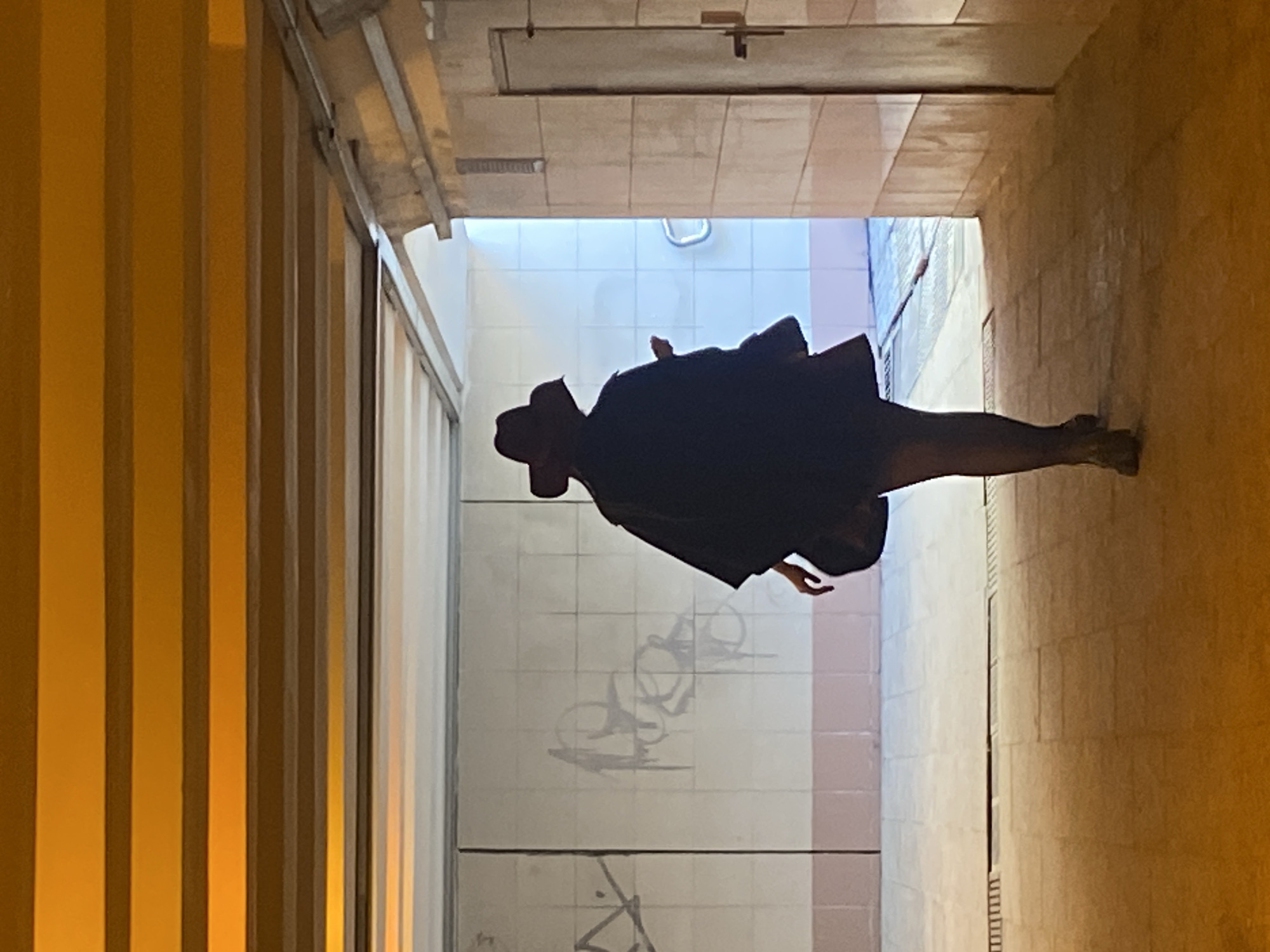 A lone individual is depicted from behind, walking away from the viewer through a warmly lit pedestrian underpass in Moscow, Russia. The person wears a dark, wide-brimmed fedora-style hat, a dark, flowing poncho-style outer garment, and dark hosiery, with dark heeled footwear. A dark bag is visible on their left side, and their right arm is slightly raised.
The underpass ceiling is constructed from parallel horizontal beams, illuminated by unseen light sources casting an amber glow. The left wall is faced with light-colored square tiles, accented by a horizontal strip of pinkish-brown tiles at the base, and displays dark graffiti markings. The right wall is clad in rectangular beige tiles, featuring a plain, light-colored door with a horizontal metal handle. The floor is paved with light-toned rectangular tiles or flagstones, interrupted by a linear drainage grate in the mid-ground. The individual is moving towards a brighter section of the passage, where a white-tiled wall with a curved metal handrail is visible, indicating a turn or an exit point. The illumination transitions from a dominant amber in the immediate foreground to a cooler, brighter light emanating from the deeper section of the passage.