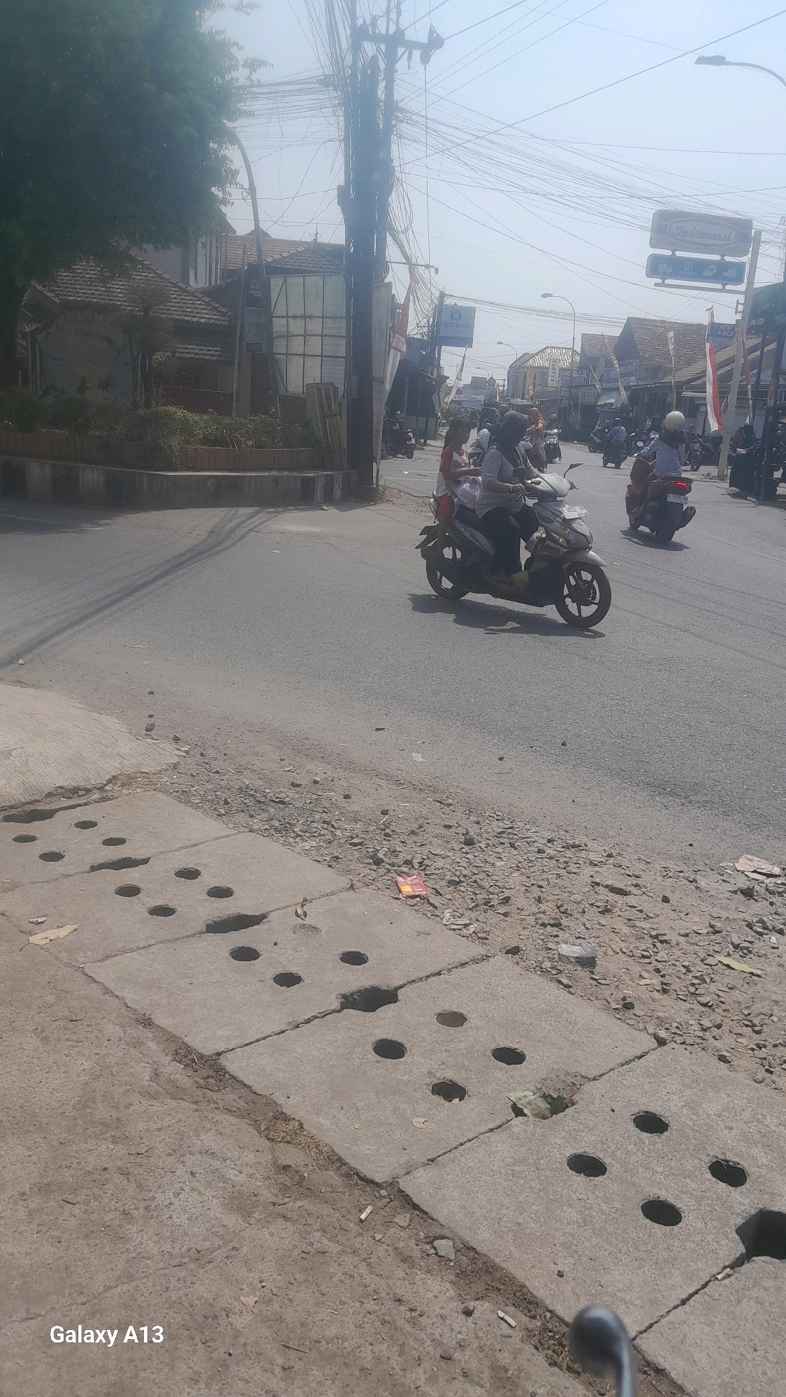 The image depicts a street scene in Kirig, Indonesia, under a bright, hazy sky. The foreground shows concrete slabs with circular openings, likely part of a drainage system, bordering a dirt and gravel shoulder. The road surface is asphalt, with tire tracks visible.
In the middle ground, several people are riding motorcycles. One scooter in the center has two occupants, a driver and a passenger, moving away from the viewer. Another motorcycle with a single rider is further down the road to the right. More motorcycles and riders are visible in the background, suggesting active traffic.
Buildings line the street on both sides. On the left, there's a low-rise structure with a tiled roof and some vegetation. On the right, various shops and businesses are apparent, with signage visible including "Indomaret," a common convenience store chain in Indonesia. A utility pole with a dense network of wires stands prominently on the left side of the road, contributing to the urban infrastructure. The overall impression is of a busy commercial street in a town or city.