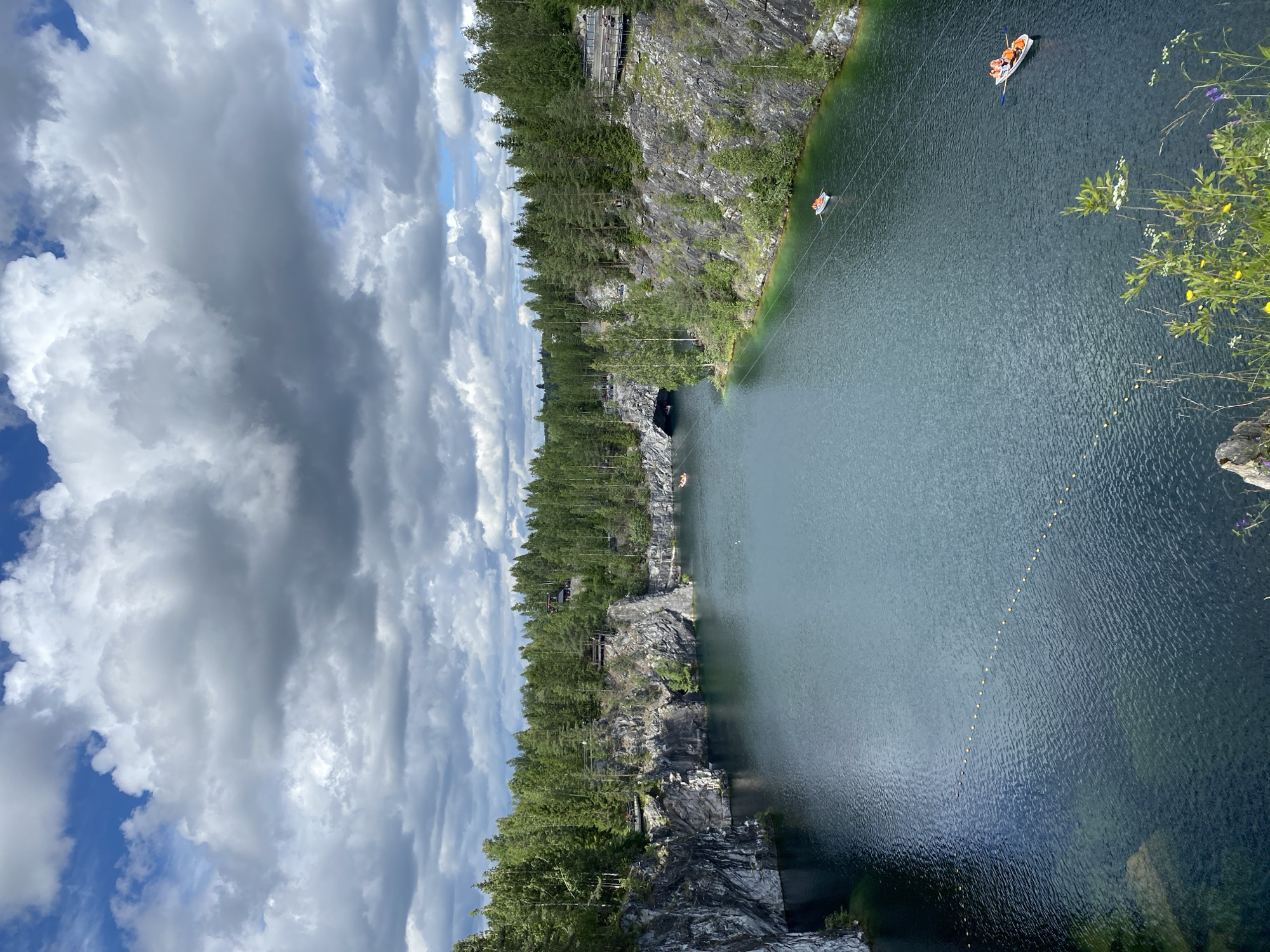 This image captures a scenic view of the Ruskeala Mountain Park in Russia. The park features a picturesque turquoise lake surrounded by steep, rocky cliffs and dense green coniferous forests. The water is calm and reflects the dramatic, cloudy sky above, which is a mix of bright white and dark grey cumulus clouds. Several small white boats with orange-vested occupants are visible on the lake, suggesting recreational activities. One boat is in the foreground, with two people visible, and another is further out in the distance. A line of yellow buoys stretches across a section of the water. Wooden walkways and observation decks are built along the cliff edges, providing vantage points for visitors. The overall scene depicts a serene natural landscape enhanced by human elements of tourism and leisure.