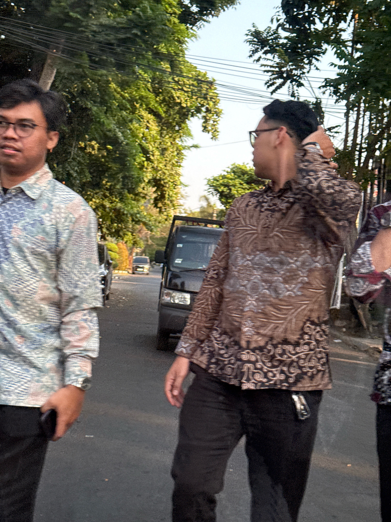 The image depicts an outdoor scene on a street, possibly in Surabaya, Indonesia, with two men walking. The man on the left is wearing glasses and a patterned shirt, with a watch on his left wrist and holding a dark smartphone in his left hand. He is looking to his left. The man on the right is seen from the side, with his hand on the back of his head, wearing a brown patterned shirt and black pants. A car is visible behind the men, and a black van is also present in the background. Trees and overhead wires suggest an urban or suburban environment. The lighting indicates it is daytime.