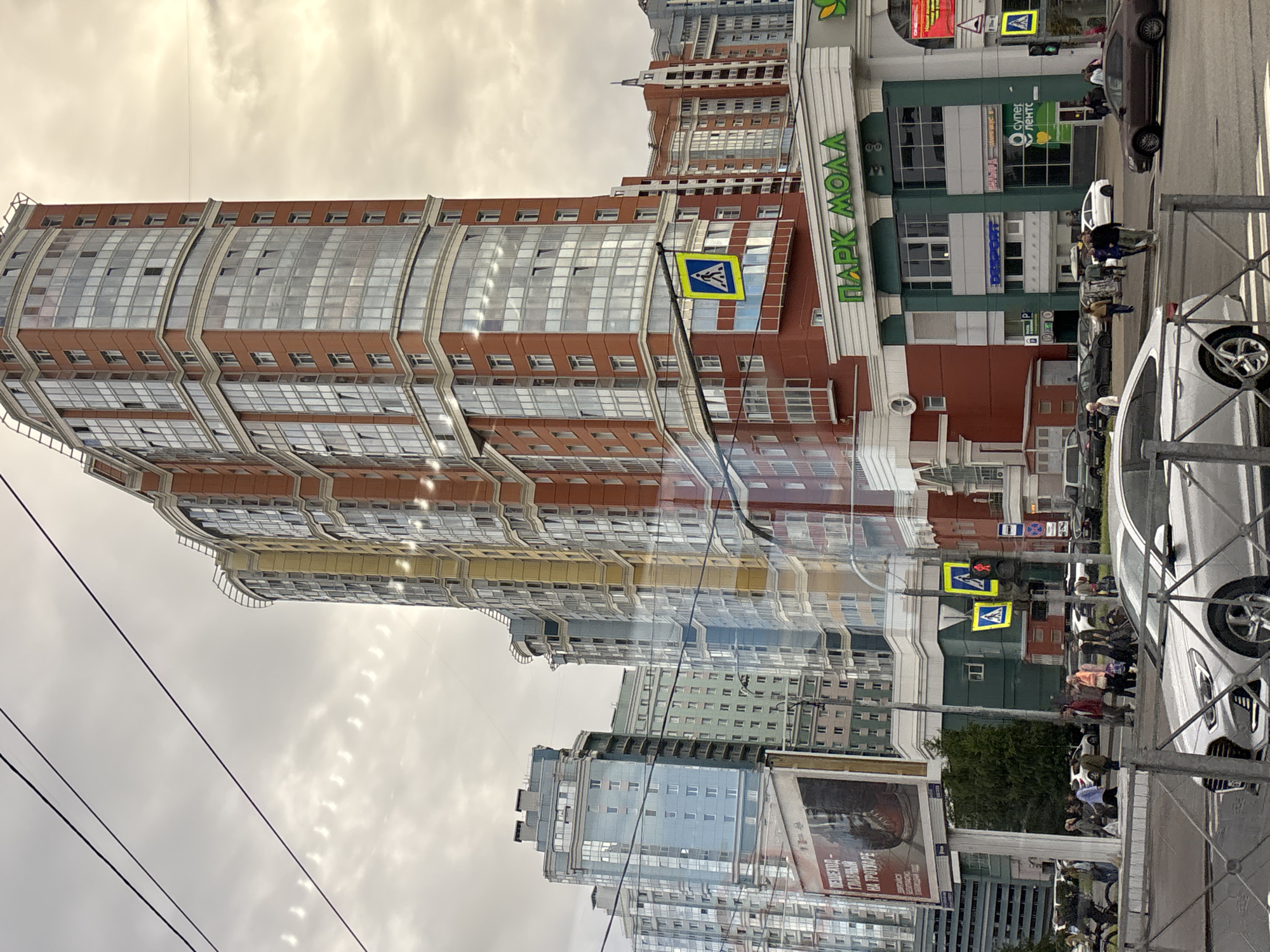 This is a vertical, eye-level, outdoor medium shot of a street scene in Sinyavino, Russia, featuring several tall, modern buildings. The sky is overcast and appears to be daytime. The buildings are a mix of residential and commercial, with prominent architectural features like curved glass facades and multiple stories. One building prominently displays the Cyrillic text "ПАРК МОЛЛА" (Park Molla), indicating a shopping center.
Several cars are visible on the street, including a white sedan and a dark-colored sedan. There are also pedestrians on the sidewalk, some of whom appear to be walking or waiting. Traffic signals and pedestrian crossing signs are present, suggesting an active intersection. The overall scene conveys an urban environment on a cloudy day. Reflections are visible on the glass facades of the buildings, hinting at the surrounding structures and possibly the sky. The image is well-lit, despite the overcast conditions.