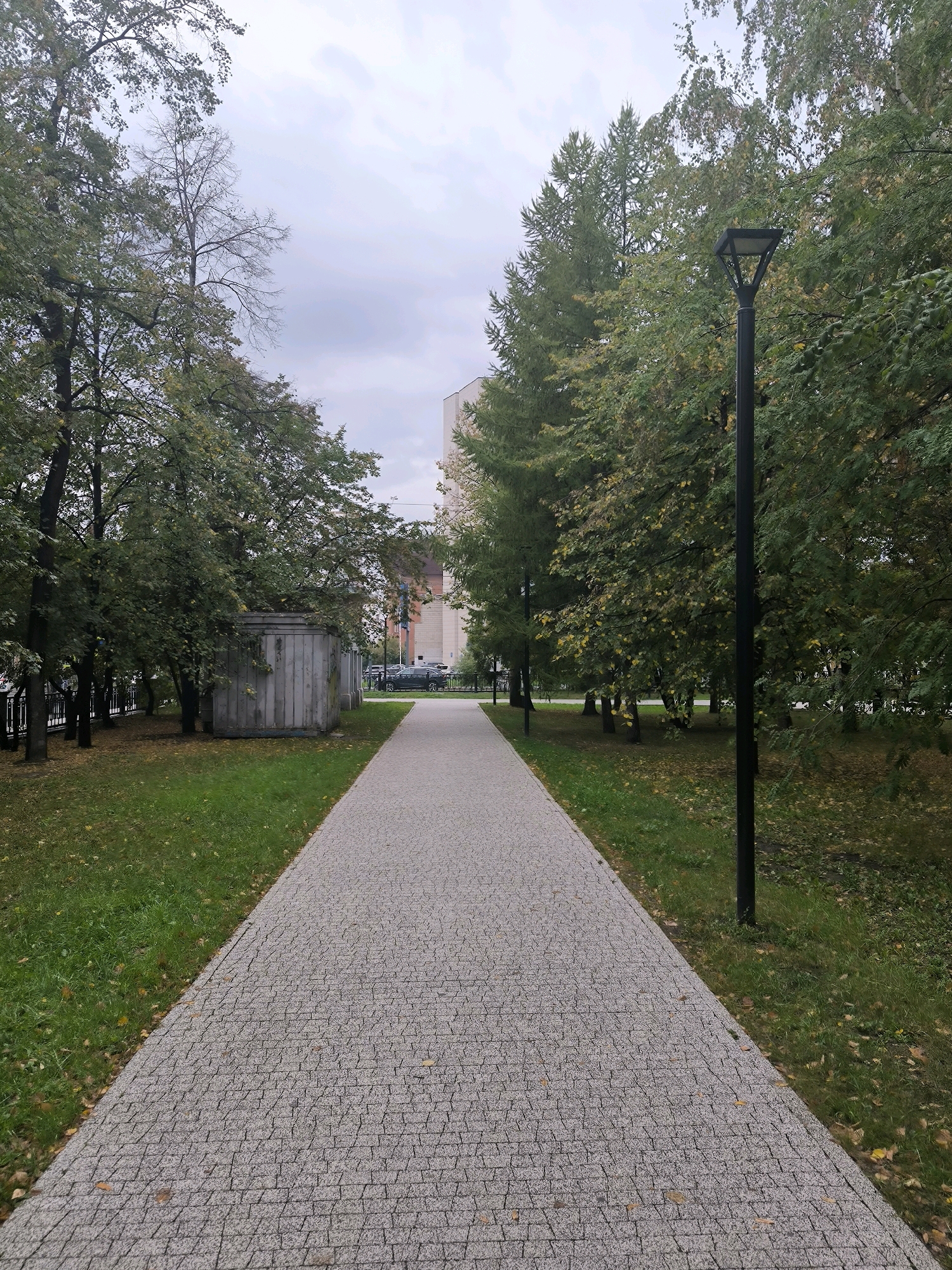 A wide, grey, stone-paved walkway runs diagonally from the foreground to the background of the image, framed by lush green grass on either side. Tall trees with dense green foliage line the walkway, creating a shaded canopy. A solitary black lamppost stands on the right side of the path. In the distance, a multi-story building with light-colored walls is visible through the trees. The sky is overcast with grey clouds. The overall scene depicts a park or public area, likely in an urban setting, during the daytime. The image is believed to be taken in Novosibirsk, Russia.