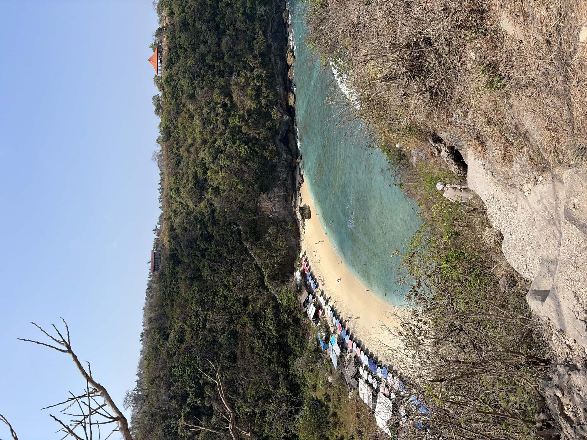 This is an aerial shot of Kelingking Beach, located in Nusa Penida, Indonesia. The beach is characterized by its white sand, turquoise waters, and lush green cliffs. Several small huts and colorful umbrellas are lined up along the shore, providing shade for visitors. In the distance, a few people can be seen swimming in the ocean and walking along the beach. The surrounding cliffs are covered in dense vegetation, with a winding path leading down to the beach. A small, orange-roofed structure is visible on top of the cliffs in the upper right quadrant of the image. The sky is clear and blue, indicating a sunny day.