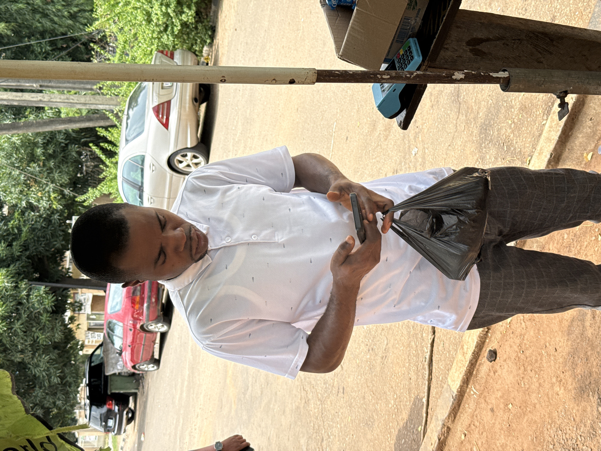 In an outdoor environment on a street in Abuja, Nigeria, a Black man stands holding a black plastic bag in his left hand and a smartphone in his right hand. He is looking down at the smartphone. The man is wearing a white polo shirt with a subtle pattern and gray checkered pants. Behind him, parked on the street, are a red sedan and a silver sedan. To the right of the man, a wooden stand is visible, with a blue card reader and a cardboard box containing items on it. A metal pole is positioned in the foreground, attached to the wooden stand. Lush green trees and bushes line the background. The ground is composed of reddish-brown soil and asphalt.
