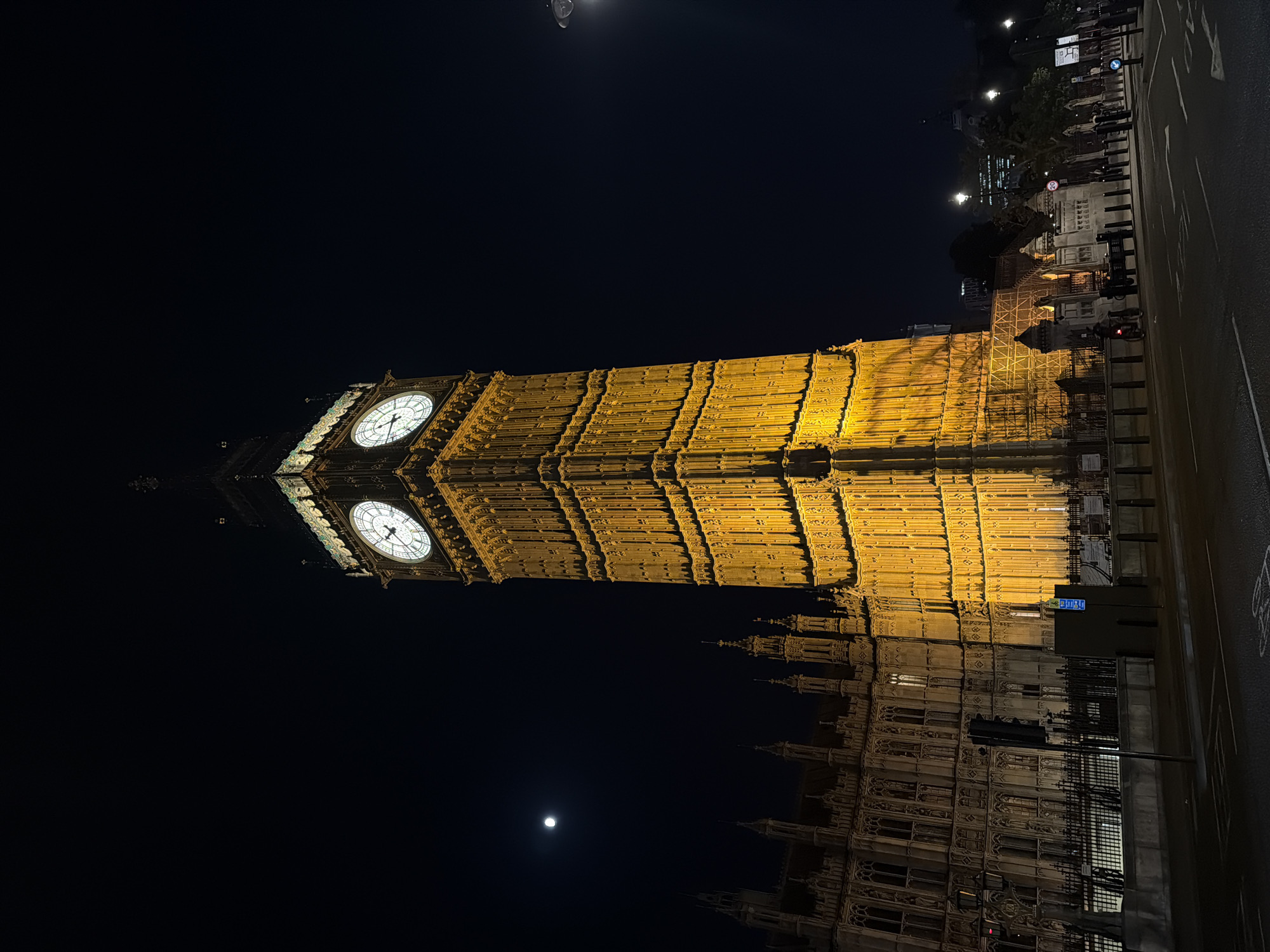 At night, a low-angle shot captures the illuminated Elizabeth Tower, known as Big Ben, in London, UK. The tower's clock faces glow brightly against the dark sky. The architectural details of the tower are highlighted in warm yellow light. Below the tower, the Houses of Parliament are partially visible. A full moon is seen in the dark sky. Along the right edge, a road with streetlights is visible, indicating an urban setting. There are no people visible in the scene.