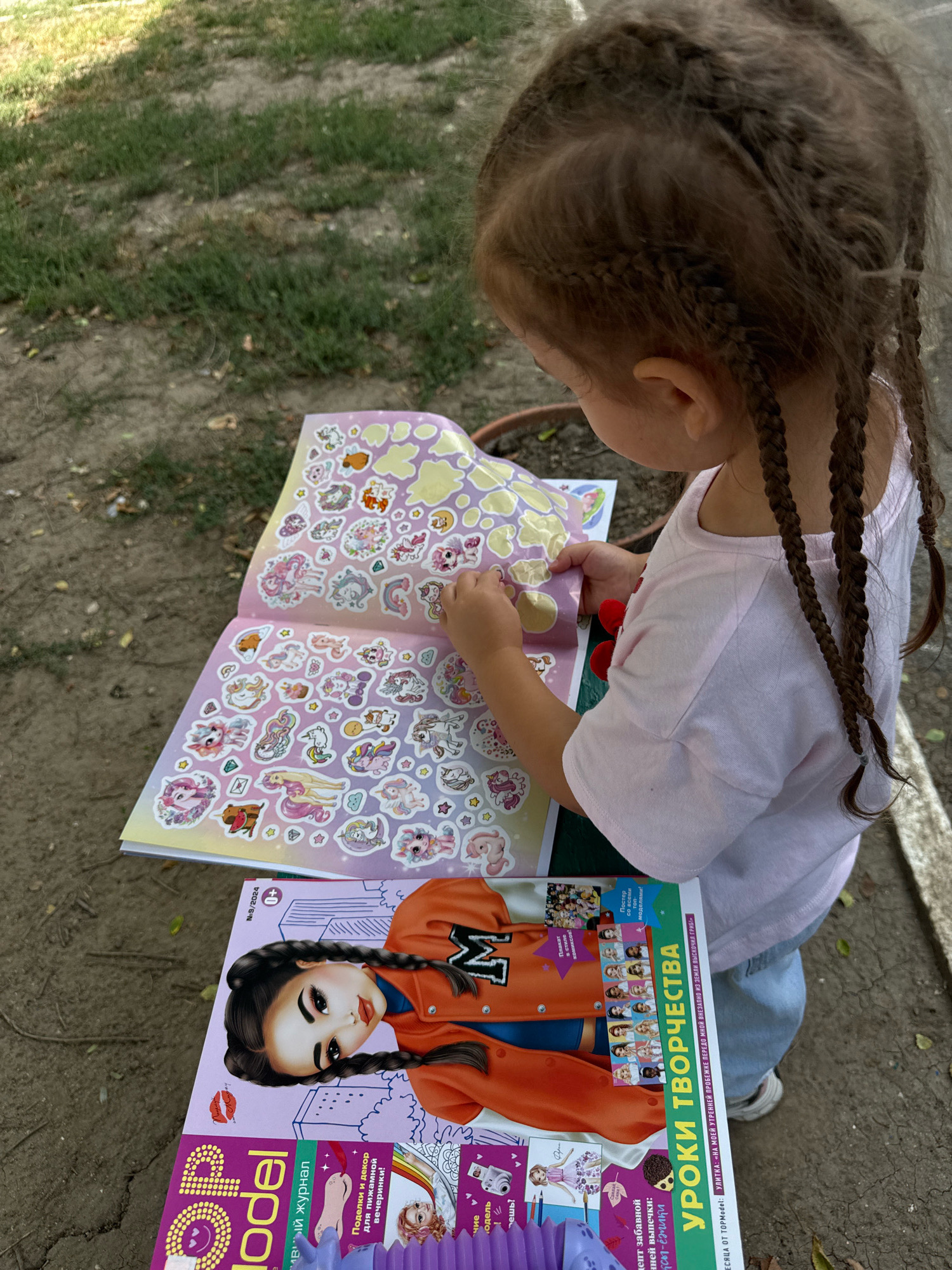 A young girl with braided brown hair is sitting outdoors, engrossed in a sticker book. She is wearing a pink t-shirt and blue jeans. The sticker book, open on her lap, is filled with colorful images of unicorns and other fantasy creatures. Beside the sticker book is a magazine titled "TopModel," featuring a stylized illustration of a young woman with braided hair on its cover. The magazine also has text in Cyrillic, including "Уроки творчества" (Lessons of Creativity).
The scene appears to be a park or a garden, with grass and dirt visible in the background, suggesting an outdoor setting on a sunny day. The girl's focused expression and the act of applying stickers indicate she is engaged in a creative and leisurely activity. The presence of the "TopModel" magazine, which is popular among young girls, suggests the girl is interested in fashion or related themes. The overall atmosphere is one of quiet concentration and playful exploration. The text on the magazine suggests a publication date of September 2024, and a location context of Chișinău, Moldova.