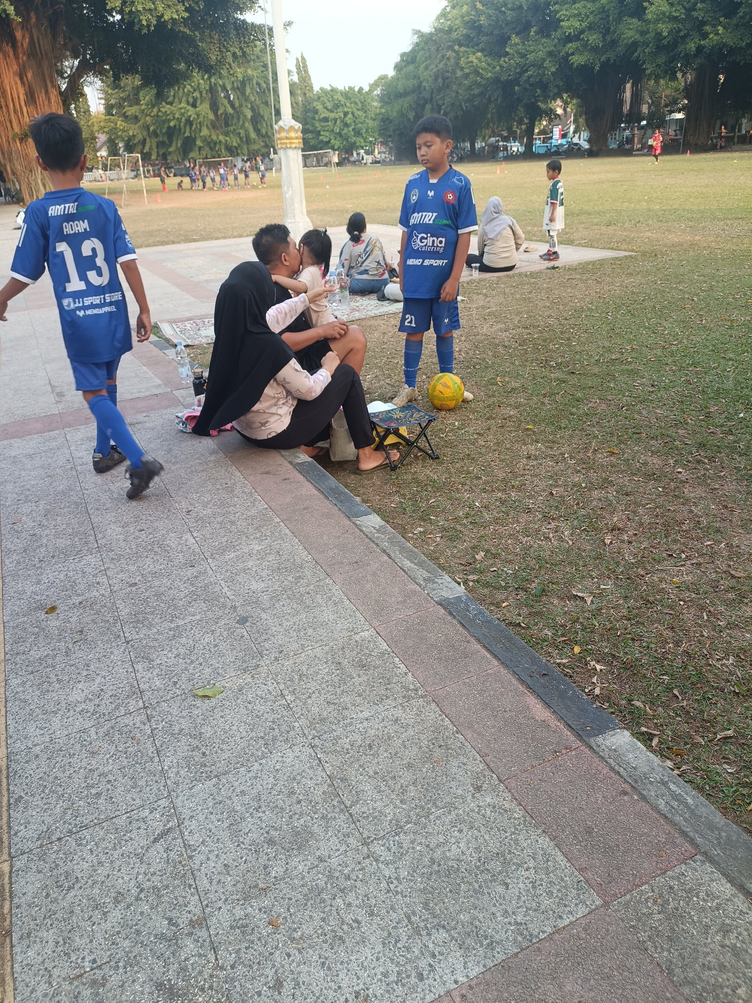 The image depicts a scene in a park, possibly Sleman, Indonesia, with several people present. In the foreground, a young boy wearing a blue soccer uniform with the number 13 on the back is walking away from the camera. To his left, a family group is seated on the ground, with a woman in a black hijab holding a child. Next to them, a young boy in a blue soccer uniform with the number 21 on his shorts stands next to a yellow soccer ball. In the background, a group of children in blue uniforms are lined up on a soccer field. The environment appears to be a grassy park with large trees and what looks like soccer goalposts in the distance. The ground in the foreground is paved with square tiles.