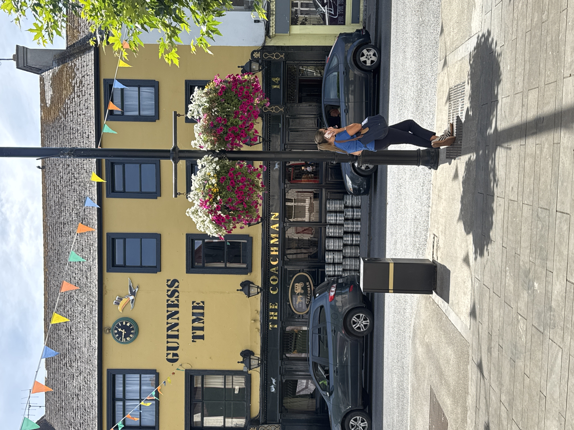 This image captures a street scene in Clonmel, Ireland, specifically focusing on a yellow building with the prominent "GUINNESS TIME" signage. The building appears to be a pub or establishment called "THE COACHMAN," as indicated by lettering above the entrance and a sign featuring a horse-drawn carriage. Suspended flower baskets, filled with pink and white blooms, hang from a lamppost in front of the building, adding a touch of color to the facade.
In the foreground, a woman with blonde hair pulled back is standing near the lamppost, engaged in a phone conversation. She is wearing a blue t-shirt, dark leggings, and beige sneakers, carrying a dark blue bag over her shoulder. To her left, a dark grey SUV is parked on the street. Behind the SUV, stacks of metal kegs are visible along the sidewalk in front of the pub.
The street is paved with asphalt, and the sidewalk is made of concrete and paving stones. Sunlight casts shadows from the lamppost and nearby trees onto the sidewalk, creating a dappled effect. Colorful triangular flags are strung across the top of the building and along the street, suggesting a festive occasion. The sky is partly cloudy.