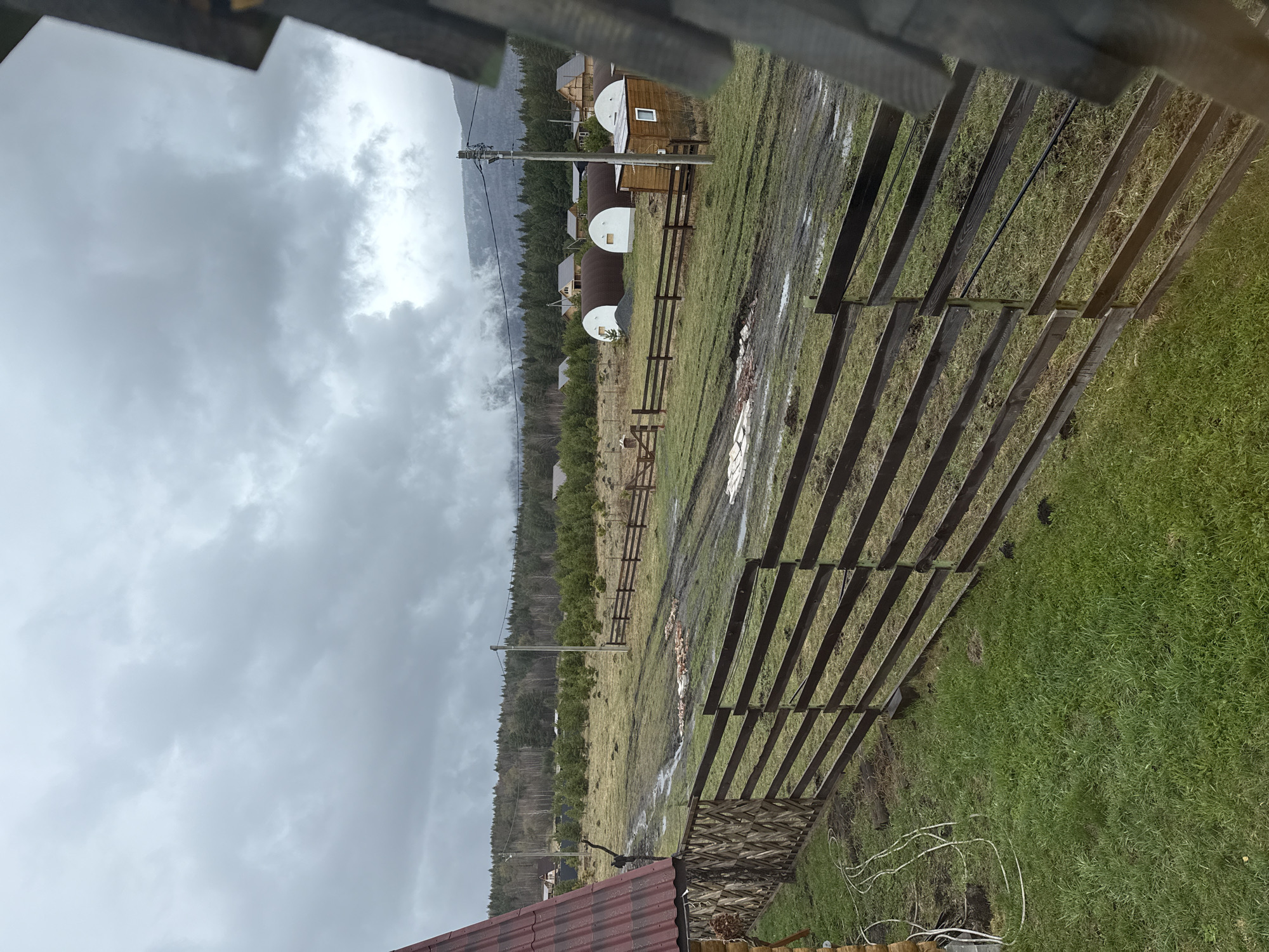 The image shows an outdoor scene in Tyulyuk, Russia, characterized by overcast, cloudy weather. A partially constructed wooden structure is in the foreground. Beyond that, the landscape includes a grassy field with patches of mud and standing water. A wooden fence runs through the field, leading towards a row of domed houses. Mountains are visible in the background, partly obscured by the low-hanging clouds. Utility poles with wires are also visible, running along the field. A rooftop is visible in the lower portion of the image.