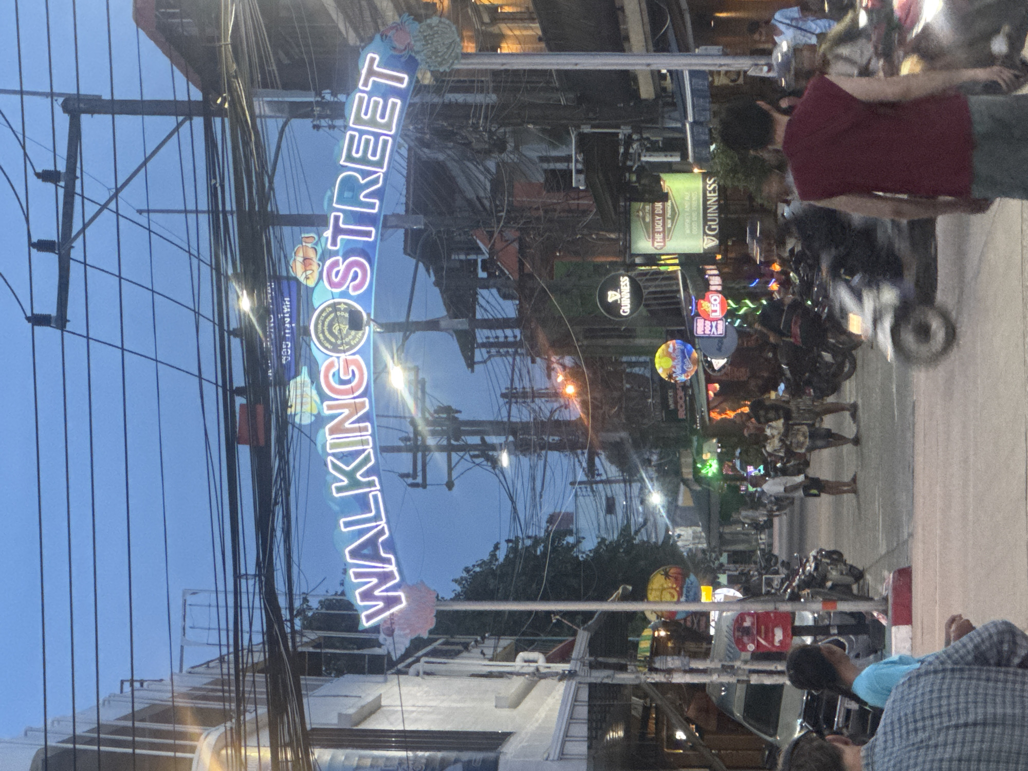 The image captures a nighttime scene on Walking Street in ตำบลเกาะพะงัน, Thailand. The "WALKING STREET" sign is prominently displayed in bright neon lettering. Overhead wires crisscross the sky above the street. Various signs for businesses, including "GUINNESS," "THE UGLY DUK," and "LEO," are visible. People walk along the street, and motorcycles are parked or moving in the vicinity. A person in a red shirt is partially visible on the right side of the image. People are sitting and appear to be engaging in conversation on the bottom right corner of the image. The environment suggests a busy commercial area, with illuminated signs contributing to the atmosphere.