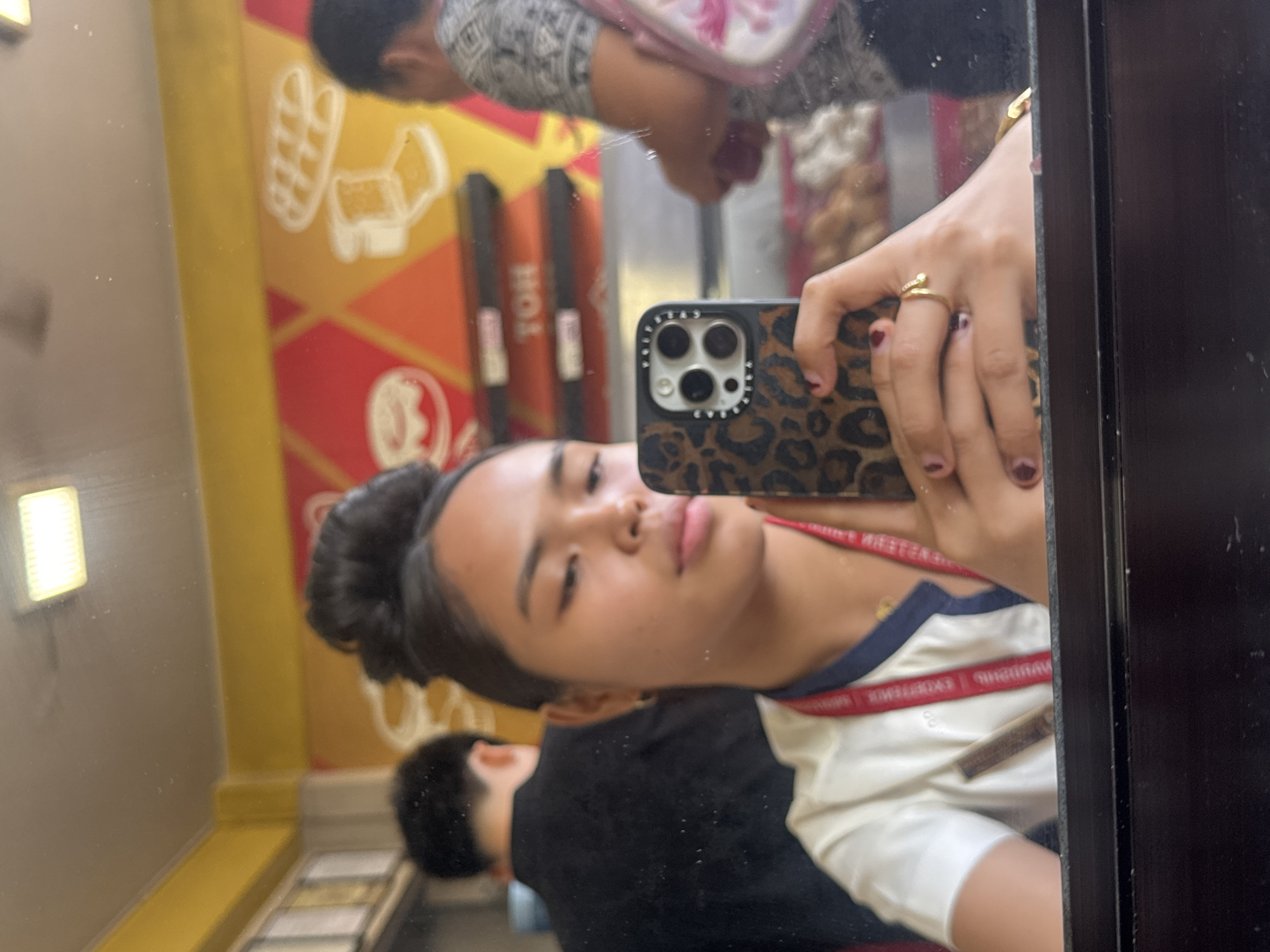 The image is a mirror selfie taken inside a food establishment in Davao City, Philippines. The foreground features a young woman with her hair in a bun, holding a leopard-print phone case. She is wearing a white shirt with a navy collar and a red lanyard around her neck. A ring is visible on her right hand. Reflected in the mirror behind her, two individuals are present. One is a person with dark hair wearing a black shirt, and the other, further in the background, is an adult wearing a patterned shirt and carrying a pink bag. The establishment's interior is visible in the reflection, with yellow and orange walls adorned with graphics of donuts and bread. Shelving units with food items and signage are also present. The overall impression is a casual snapshot within a bustling city eatery.