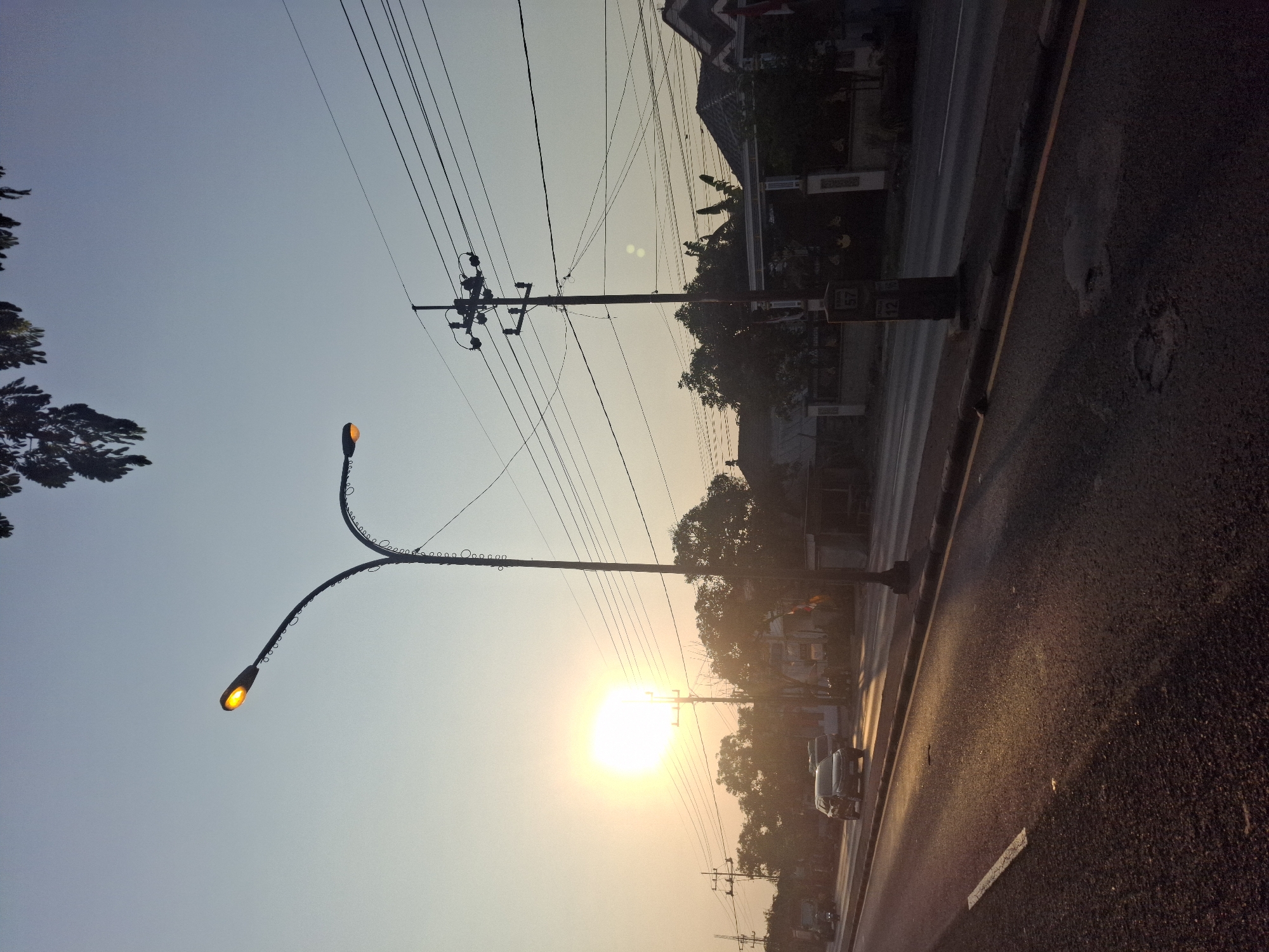 The image captures a street scene during what appears to be sunrise or sunset in Waruwetan, Indonesia. The sun is a bright, diffused orb low in the sky, casting a warm glow across the asphalt road. The road itself shows signs of wear, with some potholes visible. A white line marks the edge of a lane.
To the left of the road, a street lamp with two arched arms, each with a lit yellow lamp, stands prominently. Further down the road, several vehicles are present, including a dark SUV in motion and a truck in the distance.
On the right side of the road, a utility pole with numerous power lines stretches across the frame. Behind the utility pole, a row of houses and buildings with tiled roofs are visible amidst lush green trees. A decorative gate or archway is also present in front of one of the properties. The sky is a pale blue, transitioning to a warmer hue near the horizon where the sun is. The overall impression is of a quiet, everyday moment in a semi-urban or suburban area.