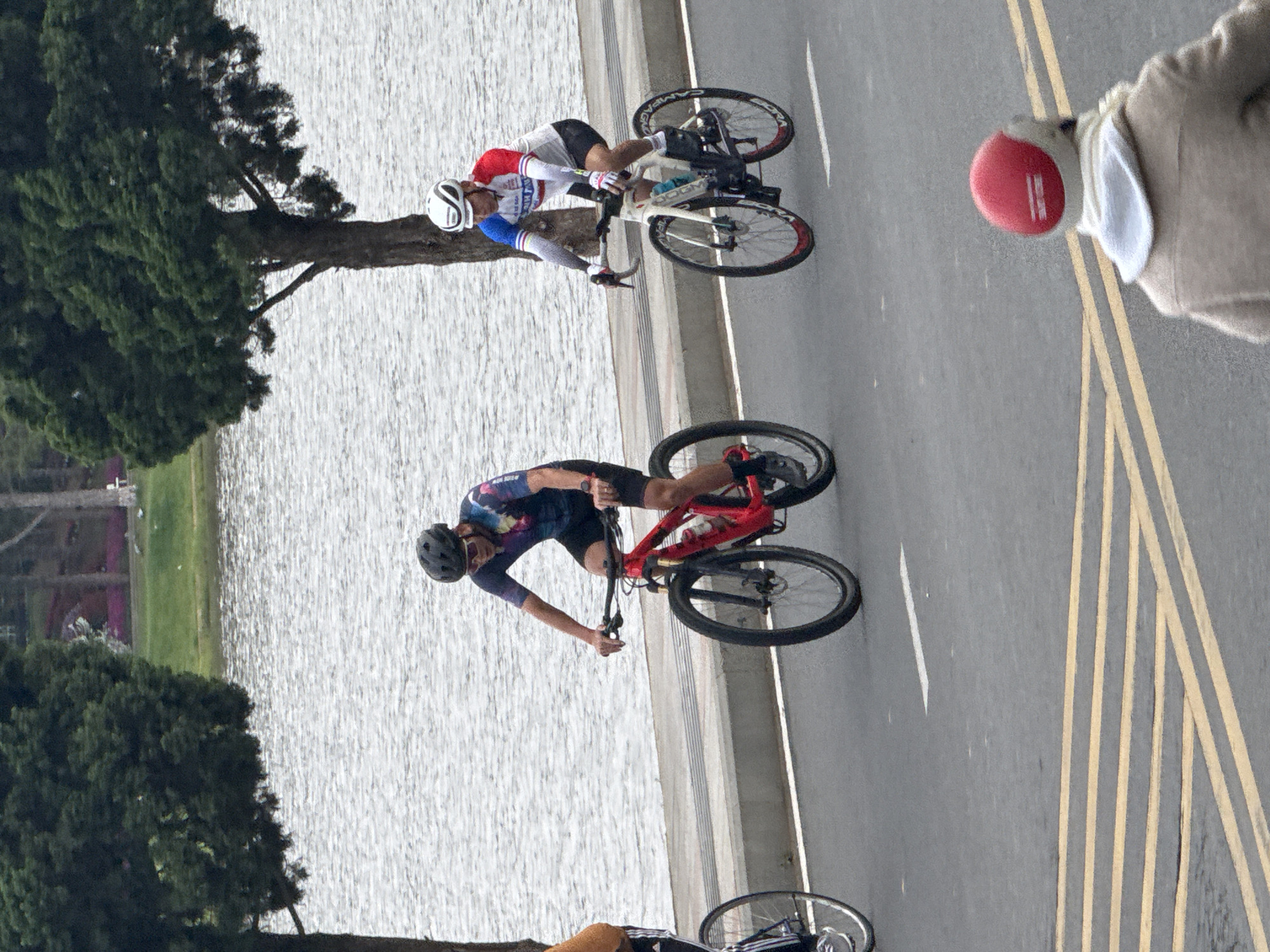 The image captures a scene on a paved road adjacent to a large body of water, likely Xuan Huong Lake in Da Lat, Vietnam, given the presence of evergreen trees typical of the region. Three cyclists are visible. In the foreground, a person wearing a red helmet and a tan jacket is riding away from the camera. In the middle ground, two cyclists are riding towards the camera. The cyclist on the left wears a dark, multi-colored cycling jersey and black shorts, riding a red mountain bike. The cyclist on the right wears a white, blue, and red cycling jersey and black shorts, riding a white road bike. The road has yellow lane markings. The water behind the cyclists appears calm with a rippled surface. Trees line the far bank of the water, and distant buildings are faintly visible. The overall atmosphere suggests an outdoor recreational activity.