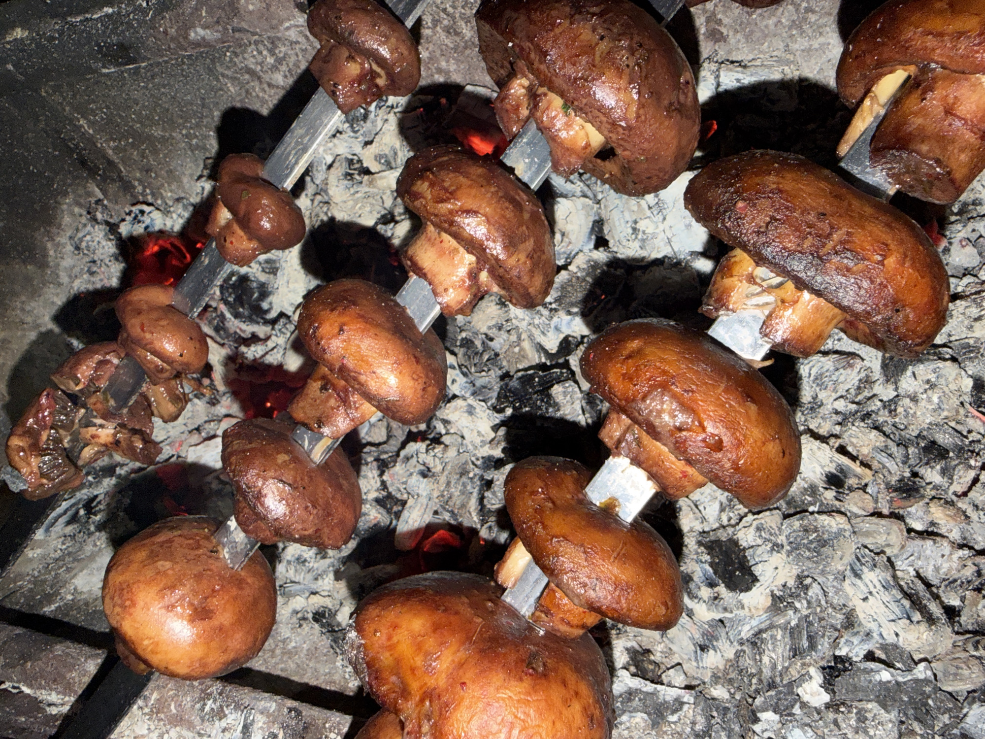 A close-up, overhead shot shows several skewers of cooked meat and mushrooms over glowing coals in a fire pit. The food is arranged on metal skewers and the mushrooms appear to be whole or halved, with the meat pieces being smaller and irregularly shaped. The scene is set in what appears to be an outdoor cooking area with visible ash and embers. The lighting is dim, with the heat from the coals creating a warm, orange glow. There are no people or identifiable landmarks visible in the image.