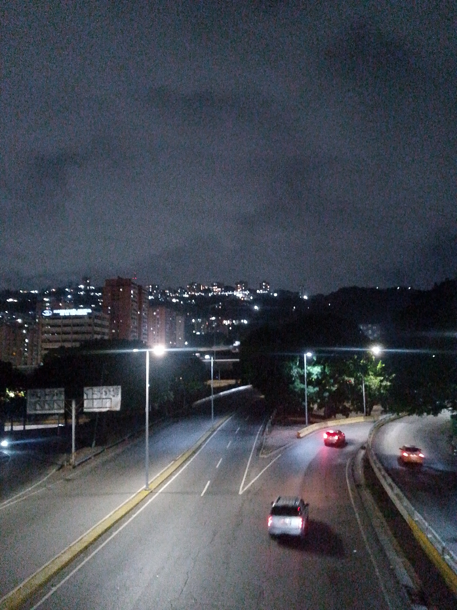 The image shows a nighttime view of a highway in Caracas, Venezuela. The highway is illuminated by streetlights, and several cars are visible. In the background, buildings and city lights can be seen on a hillside. The sky is overcast.