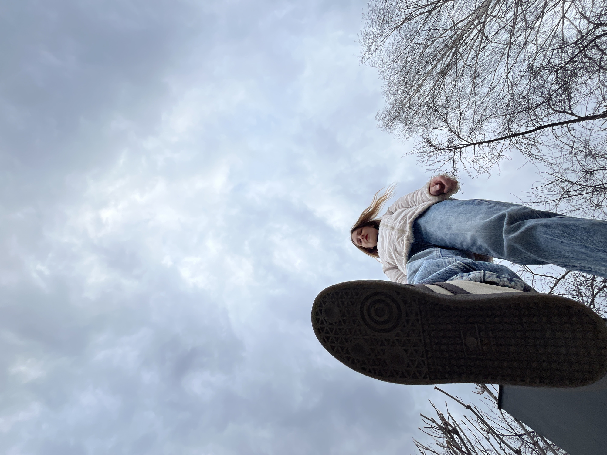 A low-angle shot from below shows a woman standing overhead. The composition features a cloudy sky filling much of the background. Bare tree branches extend from the upper right corner of the frame. The woman is wearing jeans and a white jacket. Her feet are in front, filling a good portion of the photo with the sole of a sneaker visible. The exact location is unknown. The information about Chaikovsky, Russia is not verifiable from the image.