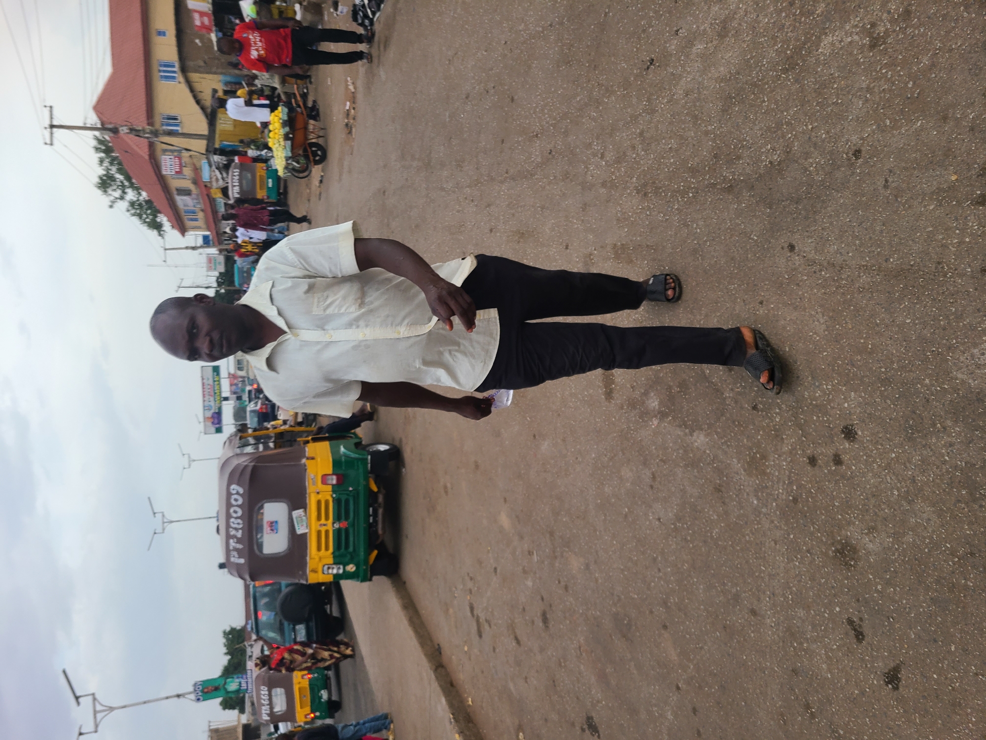 This image captures a vibrant street scene in Gura Top, Nigeria, under an overcast sky. The photograph is taken from a slightly elevated perspective looking down the street.
In the foreground, a dark-skinned man is walking on what appears to be a paved road. He is wearing a light-colored, short-sleeved button-up shirt, dark trousers, and dark sandals. He is looking towards the camera, and his left hand holds a small white object, possibly a phone or a piece of paper.
The background reveals a bustling street filled with activity. Several people are visible, some walking, others standing. Notable among the vehicles are multiple auto-rickshaws, commonly known as Keke Napep in Nigeria, which are distinctive with their green and yellow livery. One prominent auto-rickshaw has the text "PTT 28009" and "GOOD BYE!" (or possibly "GOOD LUCK!") written on its rear. Another, further down, has "PIAGGIO" visible.
Along the sides of the street, buildings with light-colored walls and reddish-brown roofs line the area, suggesting commercial or residential structures. In one section, what appears to be a street vendor's cart is laden with yellow produce, possibly plantains or bananas. Overhead utility poles and wires crisscross the sky. The ground is a rough, paved surface, indicating a well-traveled urban or semi-urban road. The overall impression is one of daily life and commerce in an active community during daylight hours.