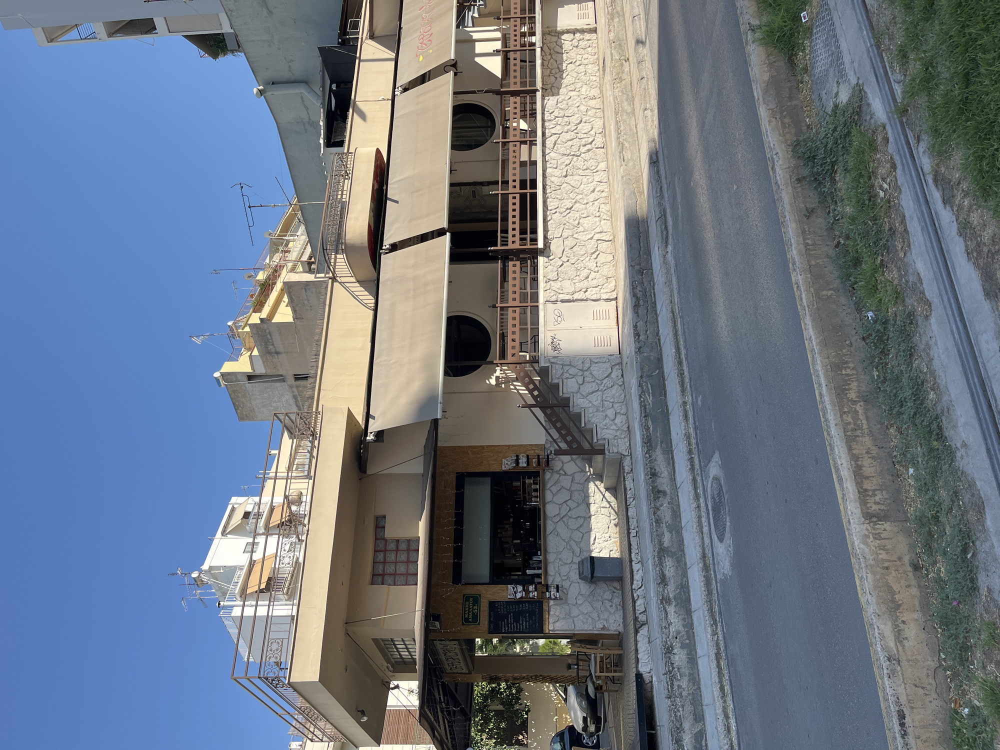 The image shows a street-level view of a multi-story building with a cafe on the ground floor in Athens, Greece. The building has a beige facade and is situated along a road with a concrete curb and a drainage channel. The cafe features a covered outdoor seating area with awnings and a stone-clad lower section. A menu board is visible outside the cafe, along with chairs and tables. The upper floors of the building have balconies and windows. The sky is clear and blue. The scene appears to be set in a sunny urban environment.