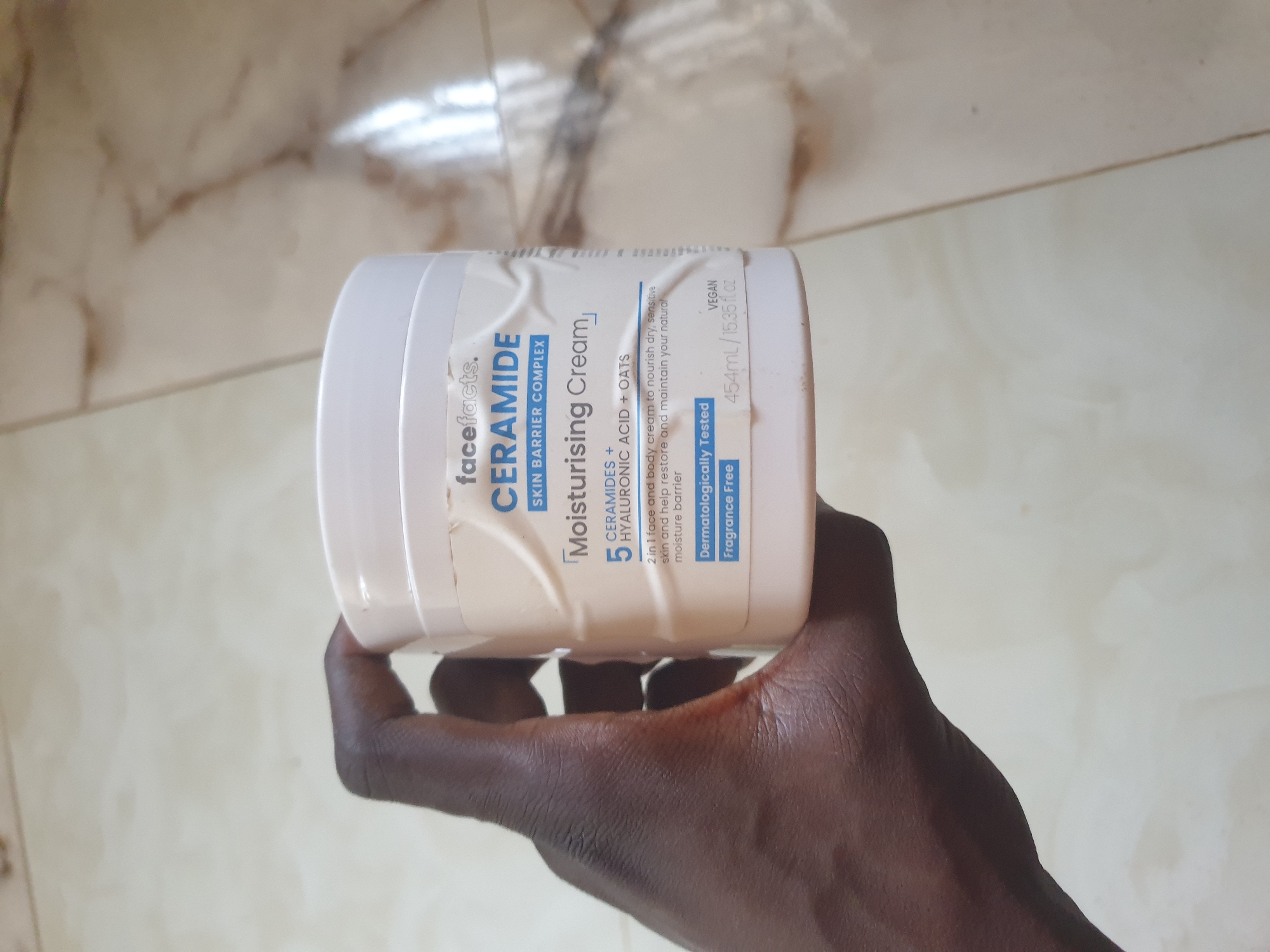 A hand, belonging to a person with dark skin, holds a white tub of "Face Facts CERAMIDE SKIN BARRIER COMPLEX Moisturising Cream". The tub is light pink and white and displays text including "CERAMIDES + HYALURONIC ACID + OATS", "2 in 1 face and body cream to nourish dry, sensitive skin and help restore and maintain your natural moisture barrier.", "Dermatologically Tested", "Fragrance Free", "VEGAN", and "454ml/15.35 fl.oz". The background appears to be a tiled floor with a marbled pattern. The image is taken from a slightly elevated angle, with the tub partially obscuring the hand. There is no discernible indication of the specific location in Anilelerin, Nigeria.