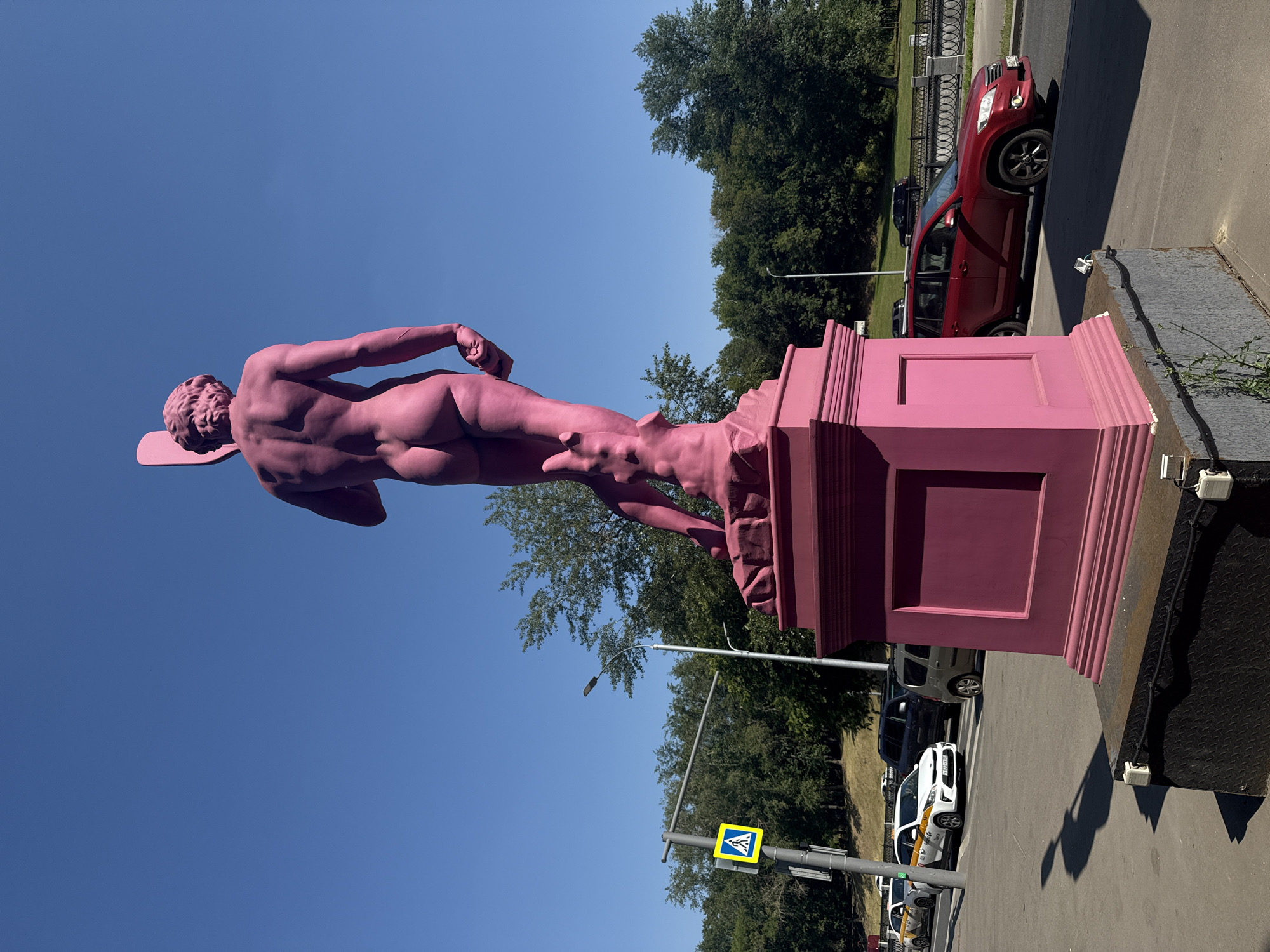 The image shows a vibrant pink statue of a male figure, viewed from the back, standing on a pink pedestal. The statue is a depiction of David and is located in the city of Zhabkino, Russia. The statue is set against a clear blue sky. In the background, there are trees, a street with cars, and a pedestrian crossing sign. One of the cars visible is a red SUV. The lighting suggests it is daytime, with the sun casting shadows.