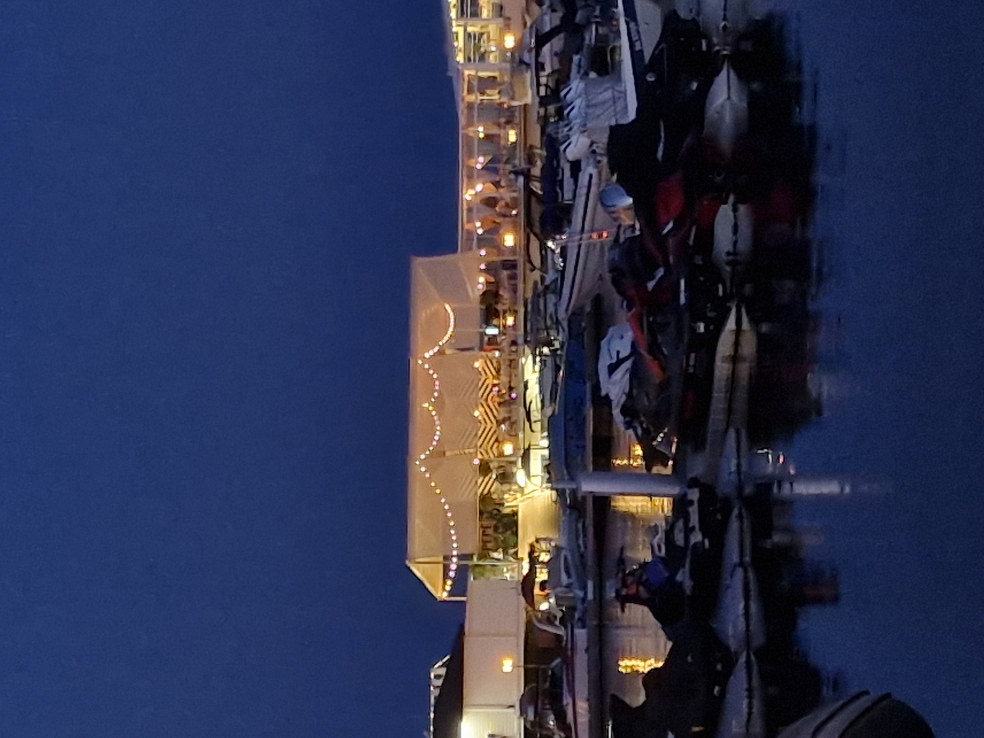 The image depicts a marina at dusk or night in Sveti Vlas, Bulgaria. The sky is a deep blue. In the foreground, several jet skis are parked on a dock, some covered with dark tarps and one with a red and black color scheme. The water is calm and reflects the lights from the marina.
In the middle ground, there are several boats moored in the water. These appear to be smaller motorboats. Beyond the boats, there is a waterfront establishment with a large, white tented roof illuminated by strings of warm-toned lights. This structure has a distinctive chevron pattern on its sides. Several people are visible sitting at tables under the tent, suggesting it is a restaurant or bar. Adjacent to this tented area, there are other covered sections of the establishment, also lit by warm lights, and more people can be seen inside and on patios. The building to the far left is a white structure with illuminated windows. The overall scene conveys a relaxed evening atmosphere at a seaside resort.