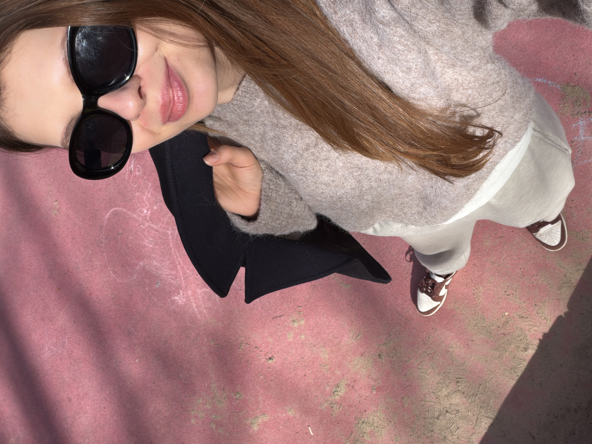 This media file presents a top-down view of a young woman standing outdoors in what appears to be a sunny urban setting in Moscow, Russia. Her face is partially visible, with large black sunglasses covering her eyes, revealing her nose and a subtle smile on her pink-tinted lips. She has long, straight brown hair that cascades over her shoulders. Her attire consists of a light beige or off-white chunky knit sweater, light-colored pants, and distinctive brown and white sneakers. She is holding a piece of black fabric, possibly a jacket, in her left hand, which is visible at the bottom of the frame. The ground beneath her is a worn, reddish-pink surface, likely concrete or asphalt, showing various scuff marks and patches of debris. Bright daylight is evident from the strong shadows cast by her body and other unseen elements onto the textured ground. The overall impression is one of a casual and relaxed moment. There is no visible text in the image.