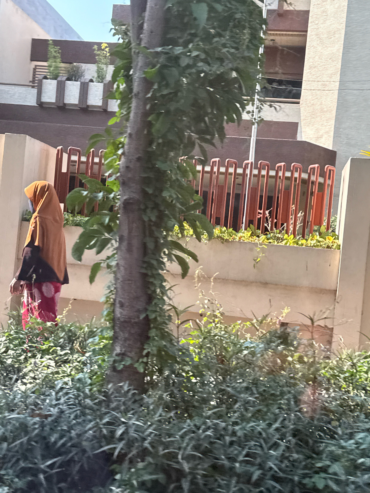 A person wearing a saffron-colored hijab and a dark top over a red and white polka-dotted skirt stands in front of a tree trunk. The person's left hand is visible. Behind the person is a building with a beige and brown facade, featuring a concrete planter box with green foliage and a railing made of vertical, orange bars. Another building with a light-colored exterior is partially visible to the right. The foreground is filled with lush green plants. The scene is bathed in sunlight, casting shadows. This image was taken in the city of Surabaya, Indonesia.