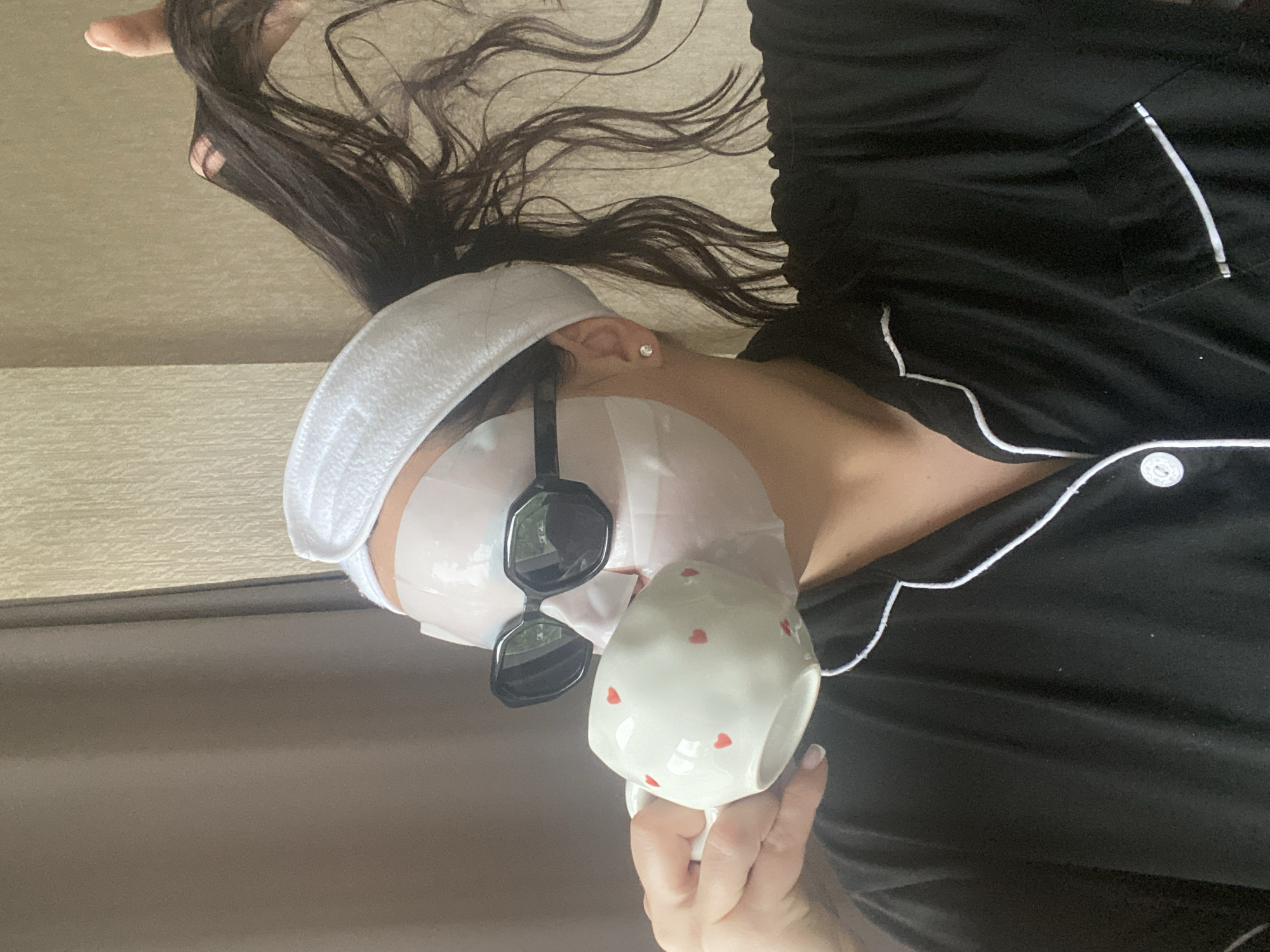 A person lies supine wearing a white towel headband, a face mask, and black sunglasses while holding a white mug with red hearts. The person is wearing black pajamas with white piping. Long dark hair fans out above the person. The location cannot be determined from the image.